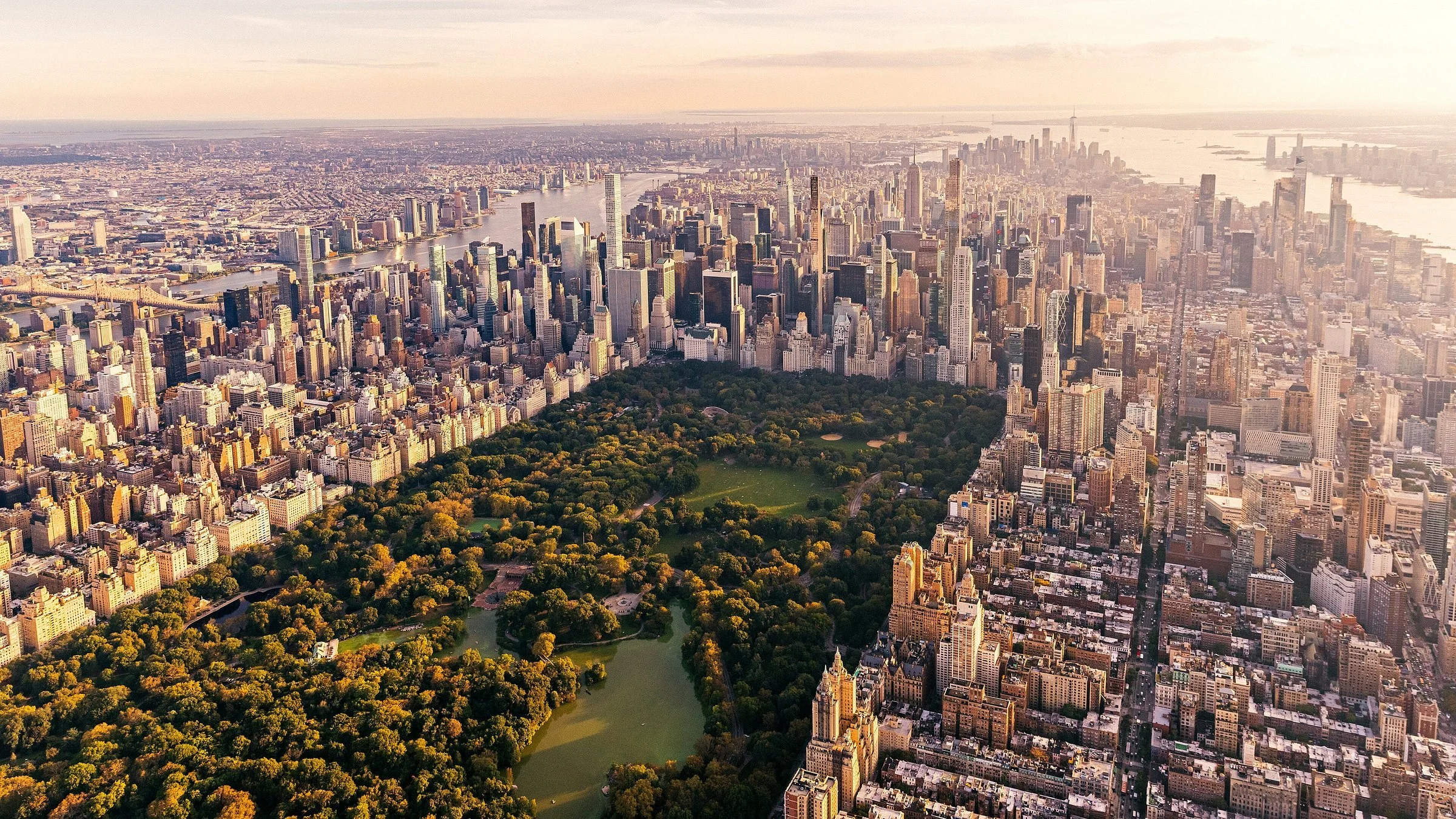 An aerial view of New York City featuring Central Park surrounded by tall skyscrapers and the East River in the background during daytime.
