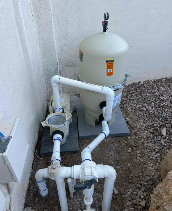 A backyard swimming pool sand filter system with white PVC pipes and valves installed on a concrete slab, next to a white wall and gravel ground.