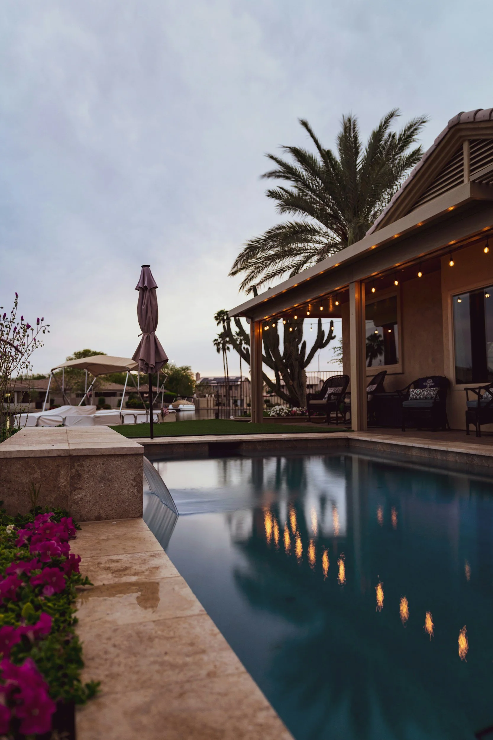 A backyard with a swimming pool, patio furniture, and palm trees at dusk. String lights are hanging from the house, and the sky is cloudy.