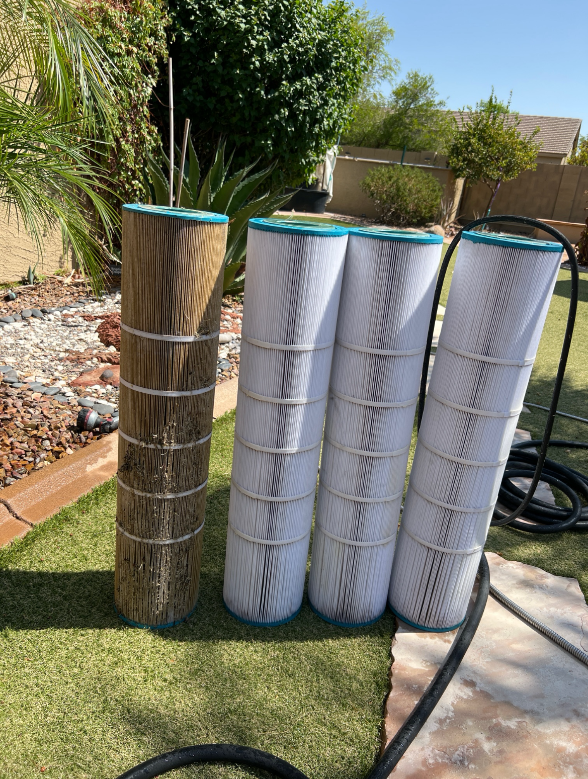 Four pool filters, three new and one used, standing upright outdoors on grass with plants and a house in the background.