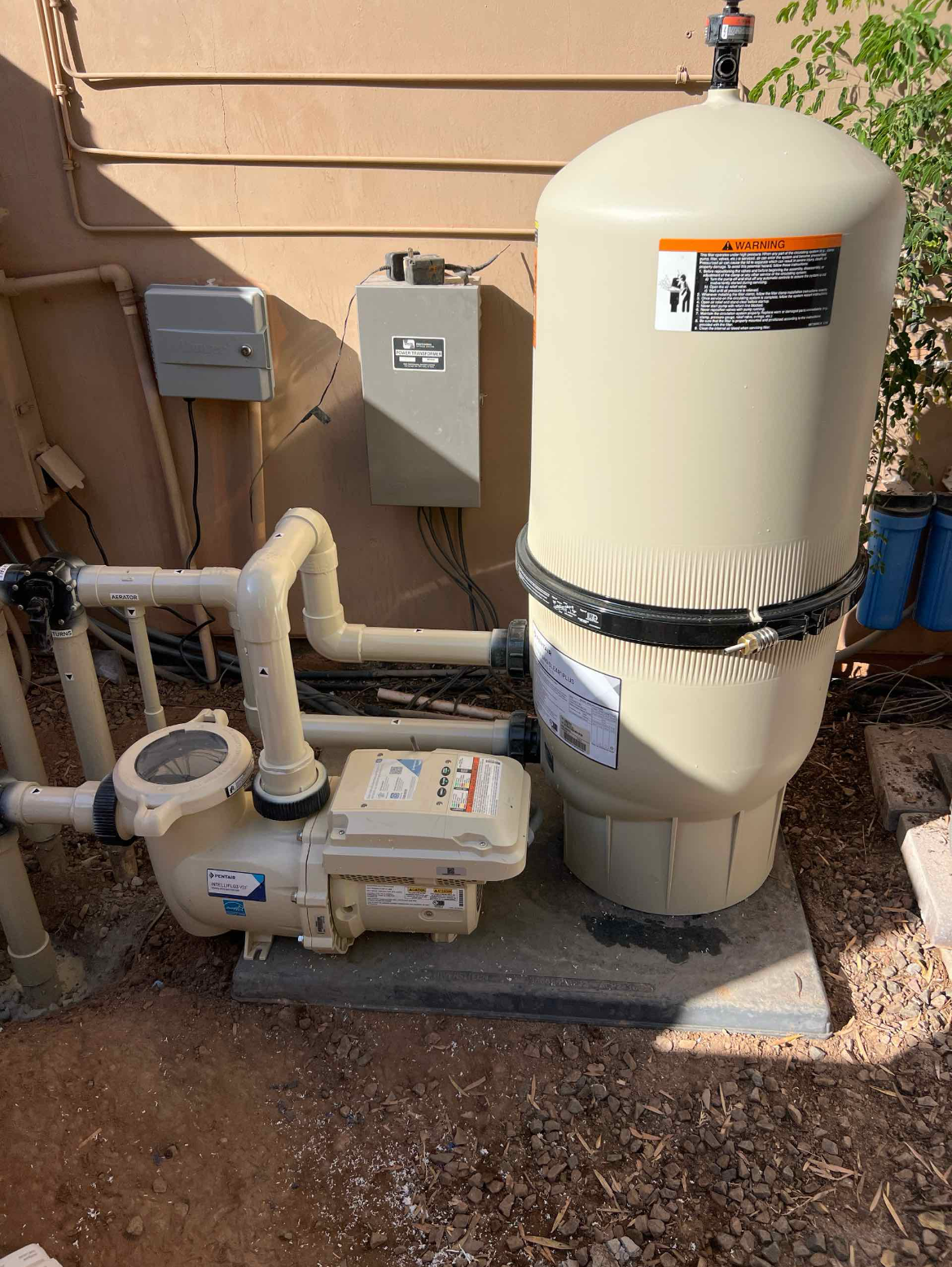 An above-ground pool sand filter system connected to plumbing and electrical components on a concrete slab outside. The system includes a large beige filter tank, a pump, and various pipes and valves, with electrical boxes mounted on a beige stucco wall.