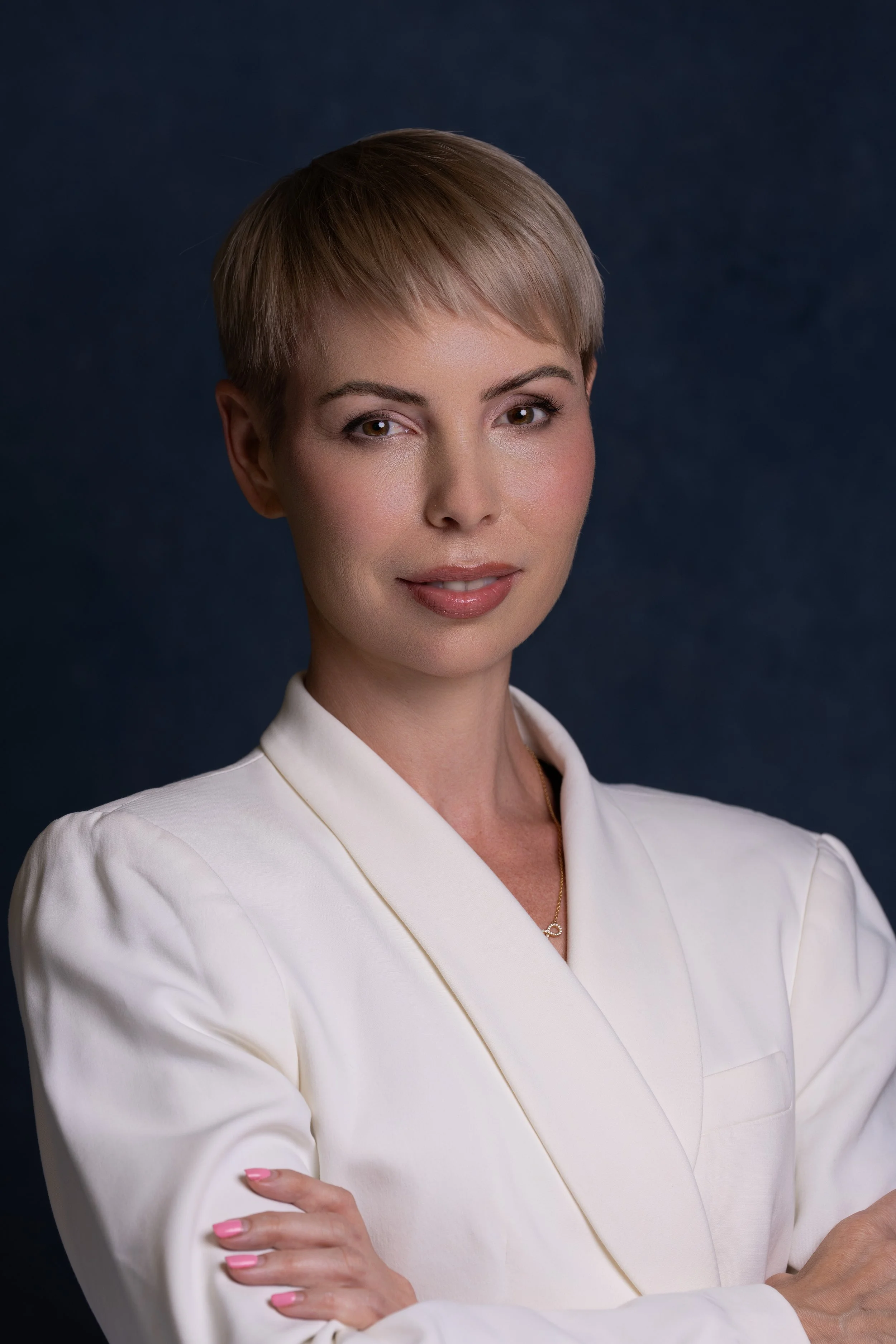 A woman with short blonde hair in a professional white blazer standing against a dark background.