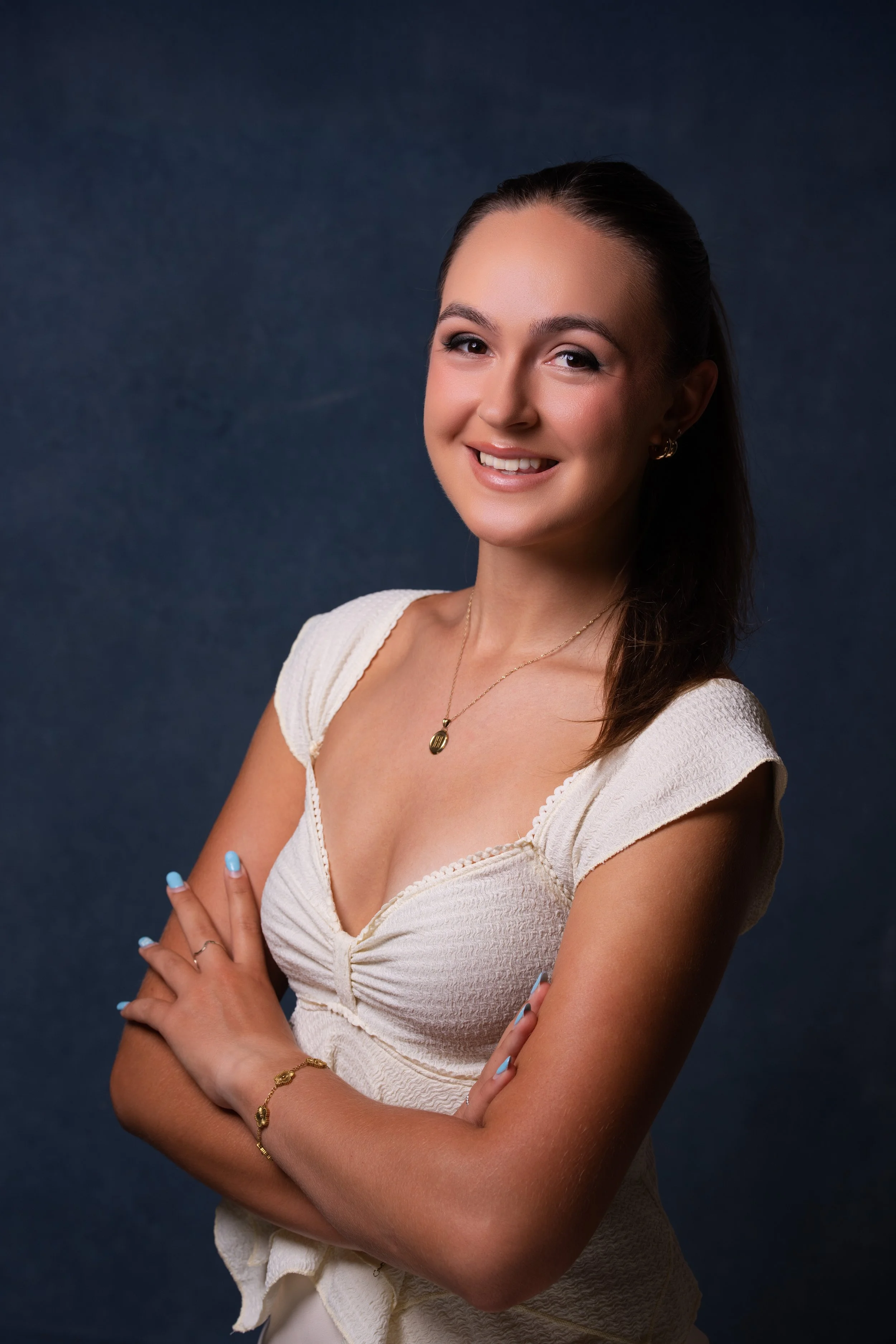 A young woman with long brown hair, wearing a cream-colored textured top, gold jewelry, and light blue nail polish, smiling with arms crossed against a dark blue background.