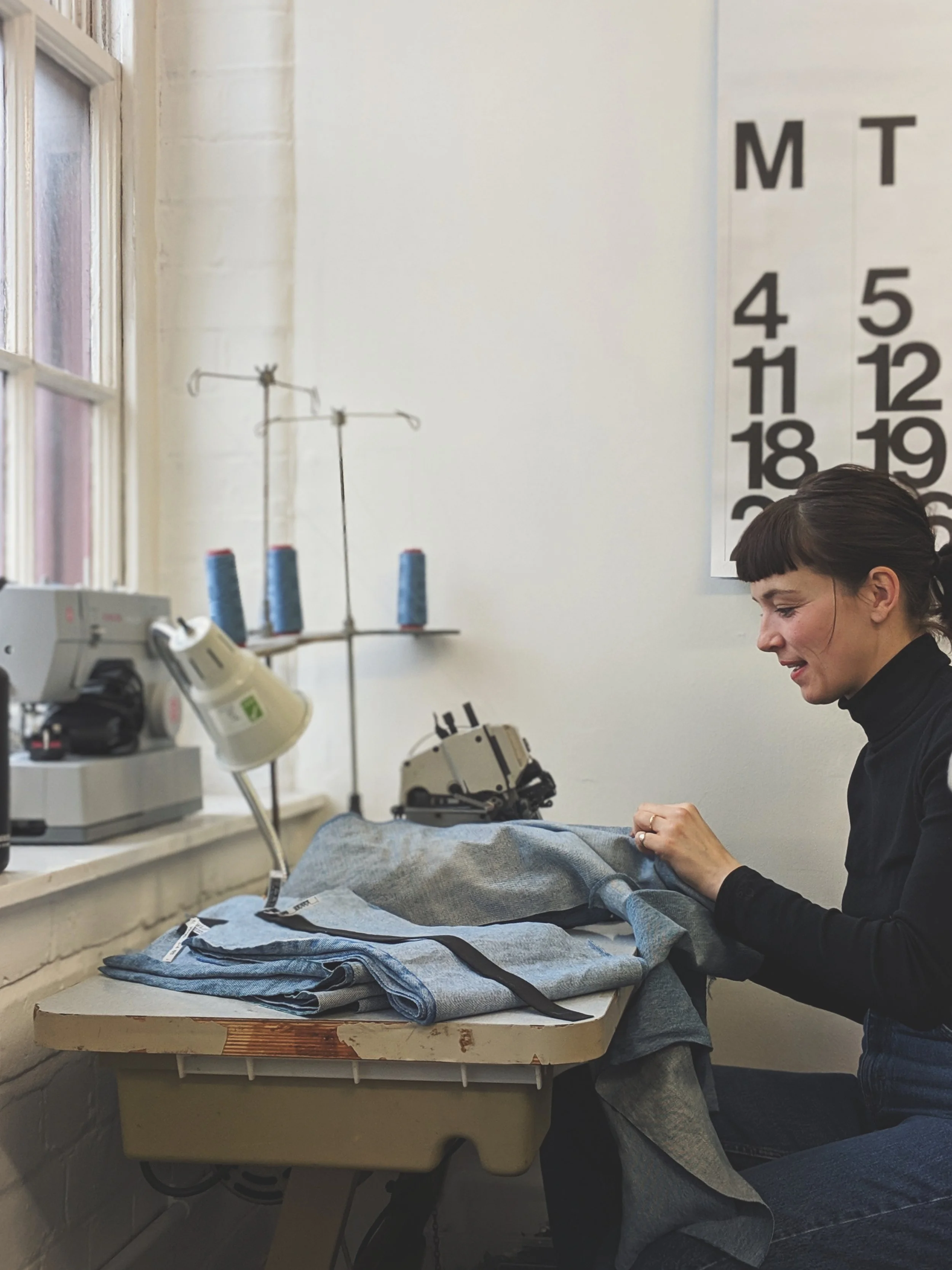 A woman with short hair smiling while sewing in a room, with a large window on the left, a row of sewing or medical tools, and a chart on the wall with the numbers 1 through 31 arranged in columns.