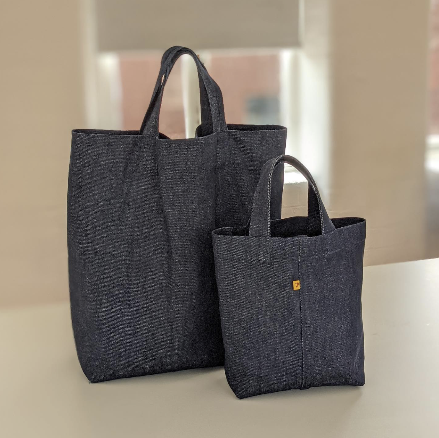 Two black fabric tote bags, one larger and one smaller, placed on a white surface near a window with a blurred outdoor background.