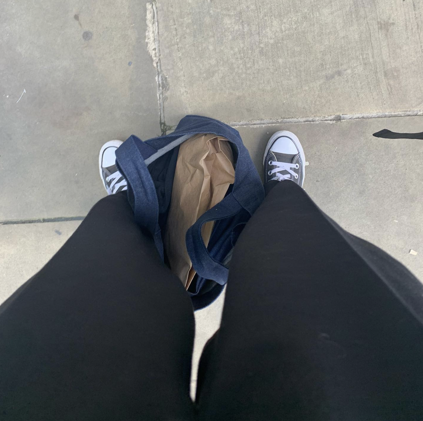 A top-down view of a person standing on a sidewalk wearing black pants and black and white sneakers, with an open blue bag containing brown paper inside.