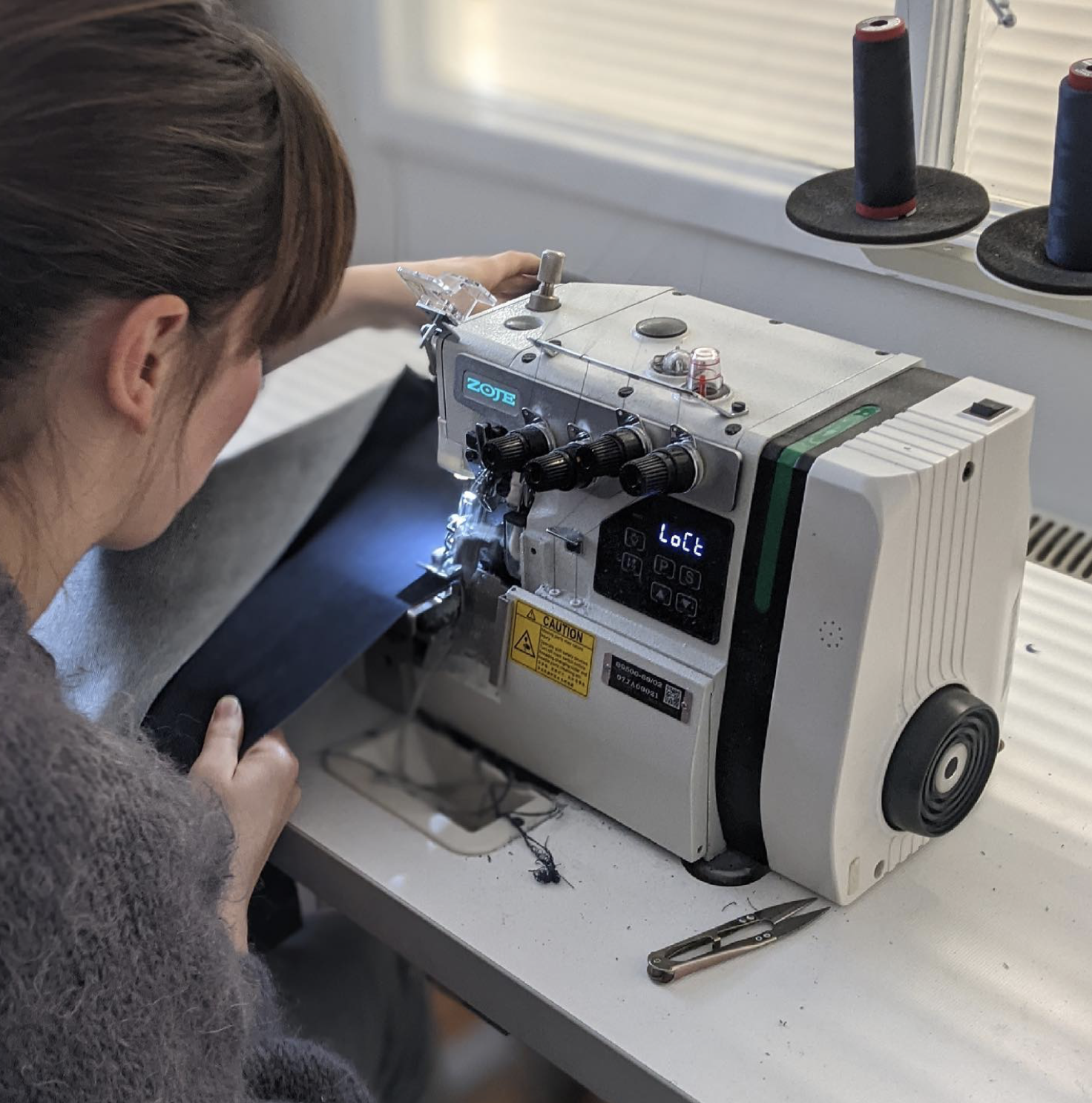 A woman working on a sewing machine, guiding black fabric through the machine in a sewing workshop.