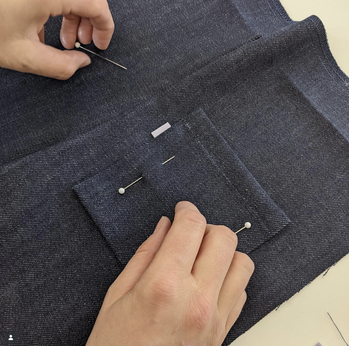 A person using a pin to sew a black pocket onto a pair of dark gray pants.