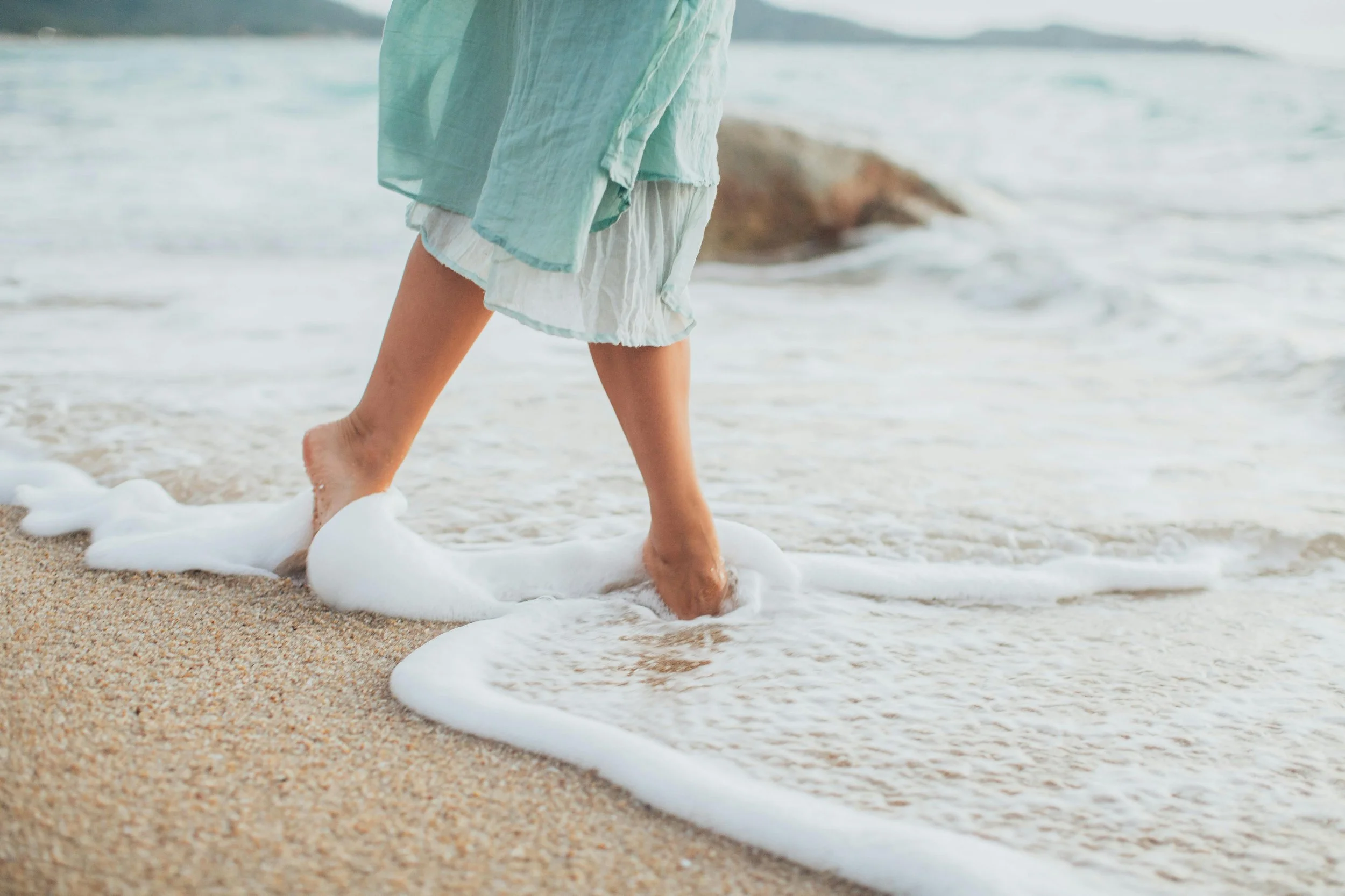 Person walking on the beach with waves and foam at their feet, wearing light-colored clothing.
