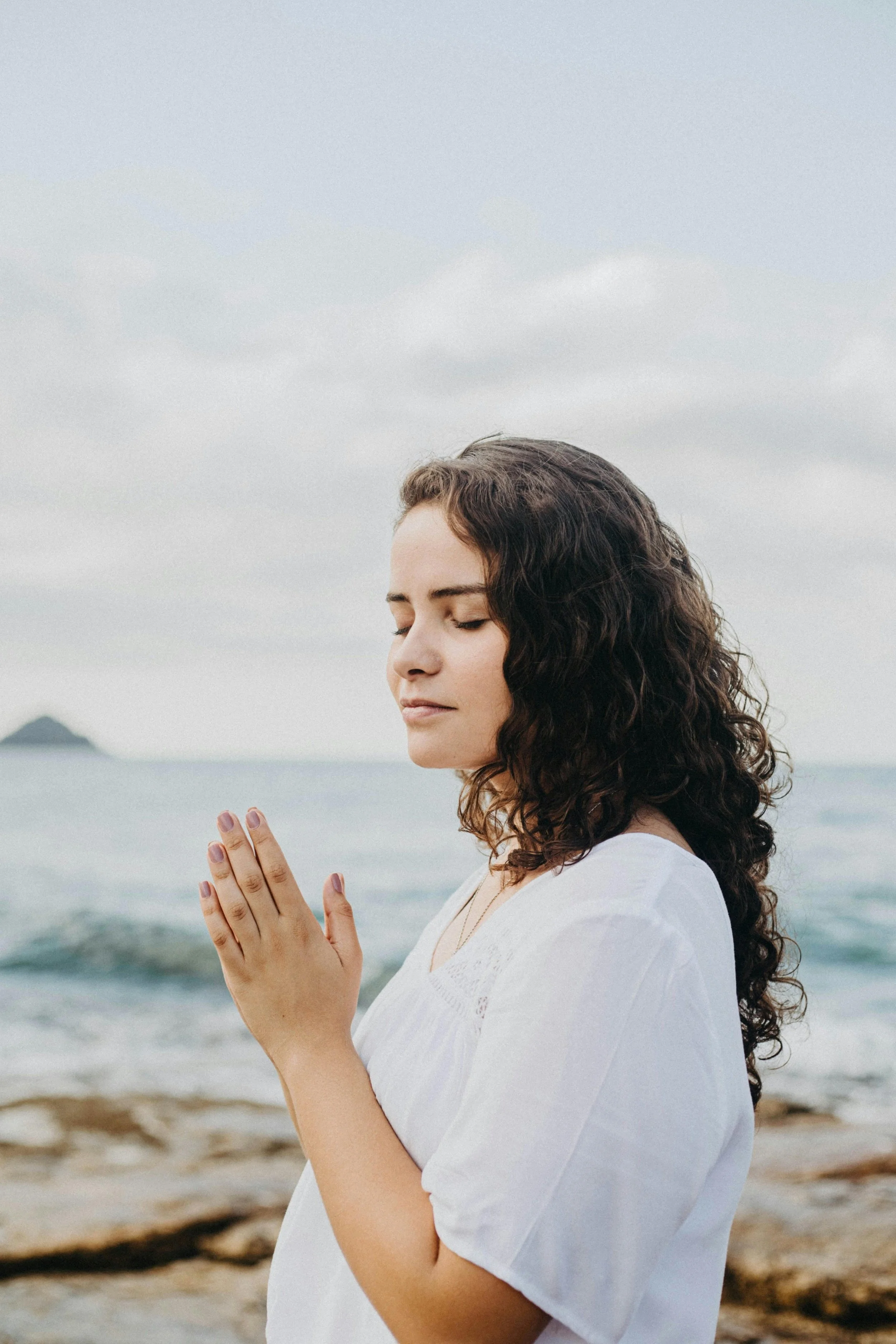A woman with curly hair and light skin standing on a beach, eyes closed, hands in prayer position, wearing a white top, with the ocean and cloudy sky in the background.