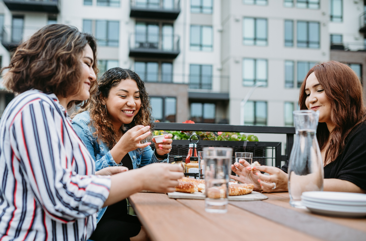 Three women sit at a table outdoors, enjoying pizza and drinks, with an apartment building in the background.