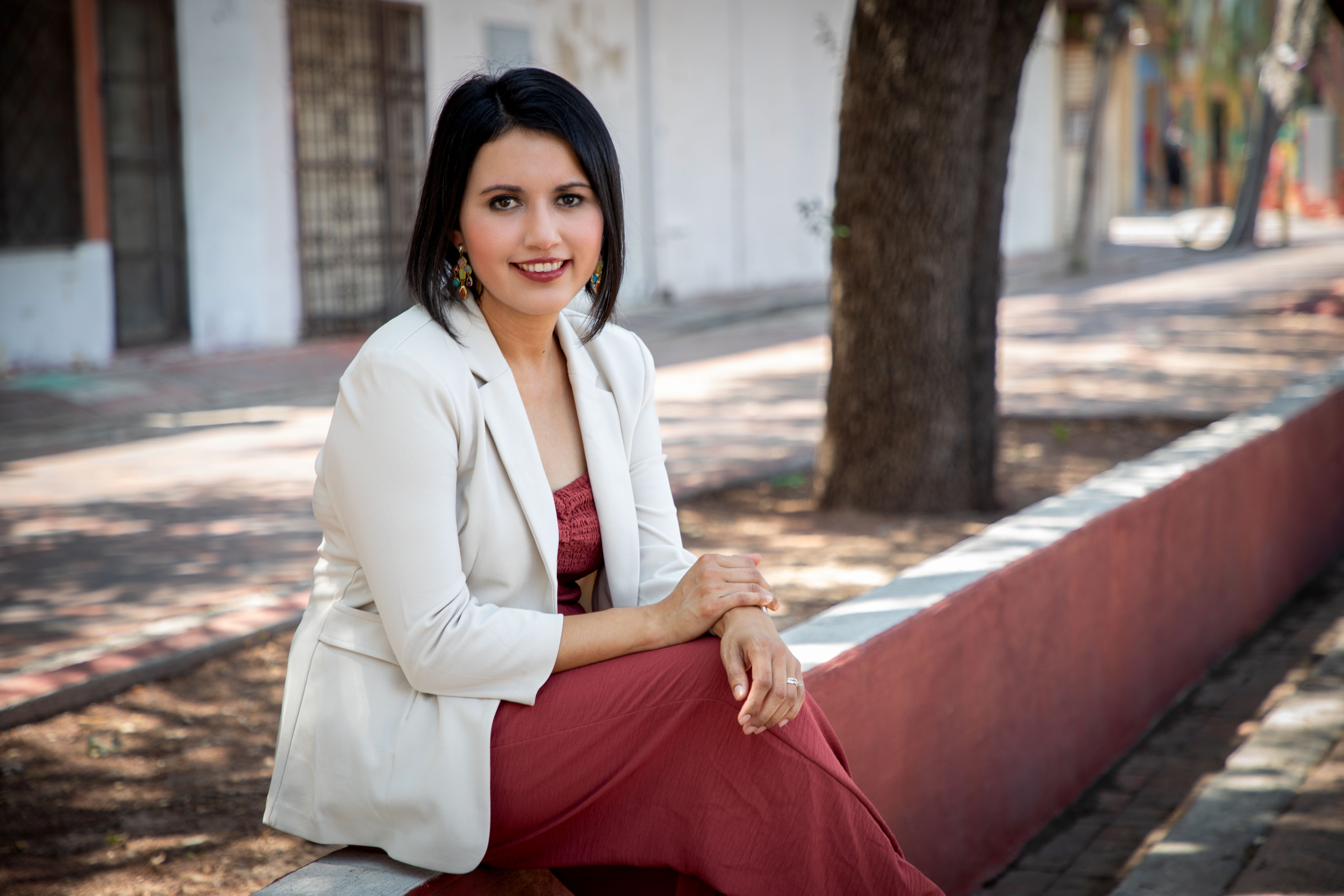 The therapist with dark hair and earrings sitting on a bench outdoors near a tree, smiling.