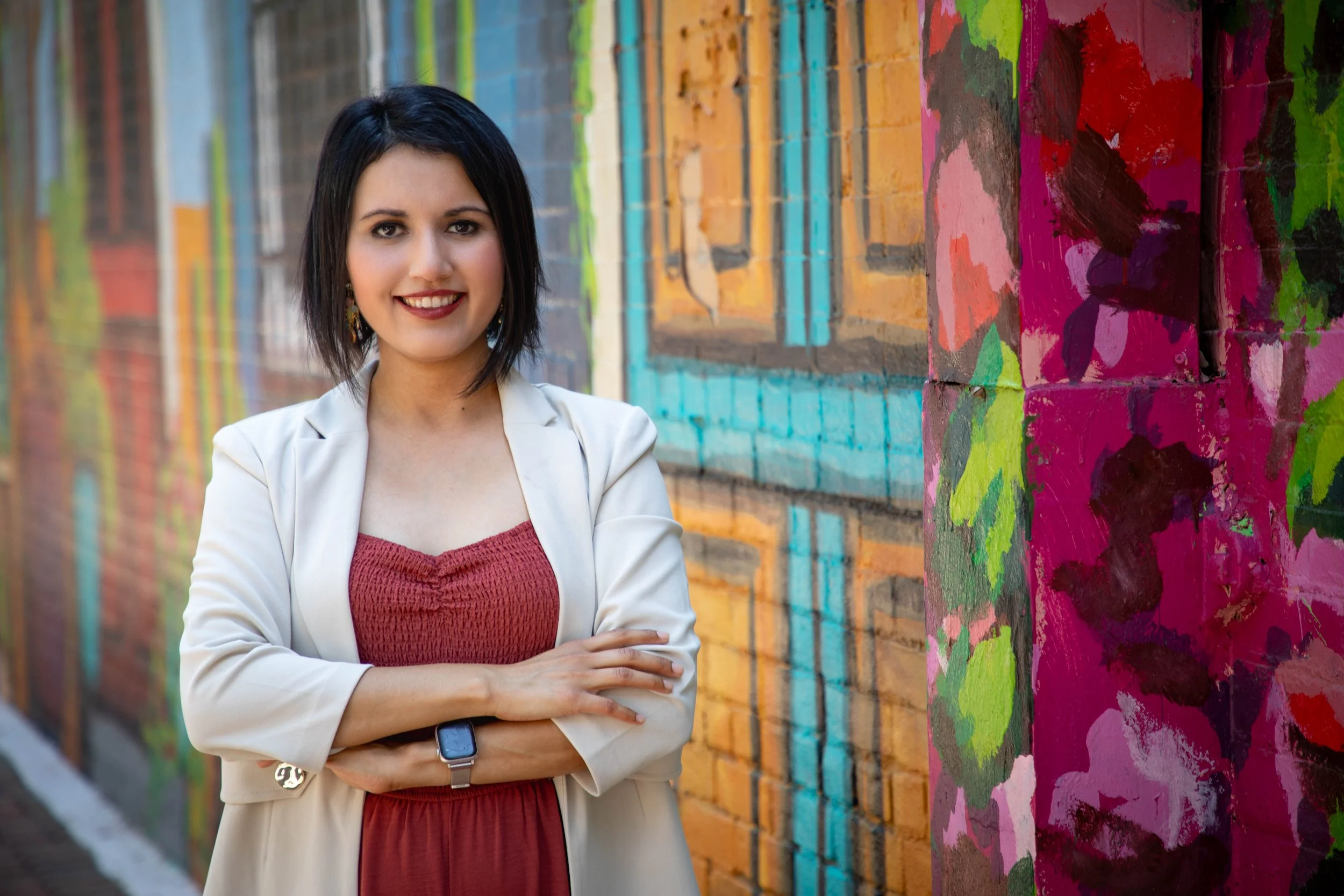 The therapist with short black hair wearing a light-colored blazer and red dress standing in front of a colorful, mural-painted brick wall with arms crossed and smiling.