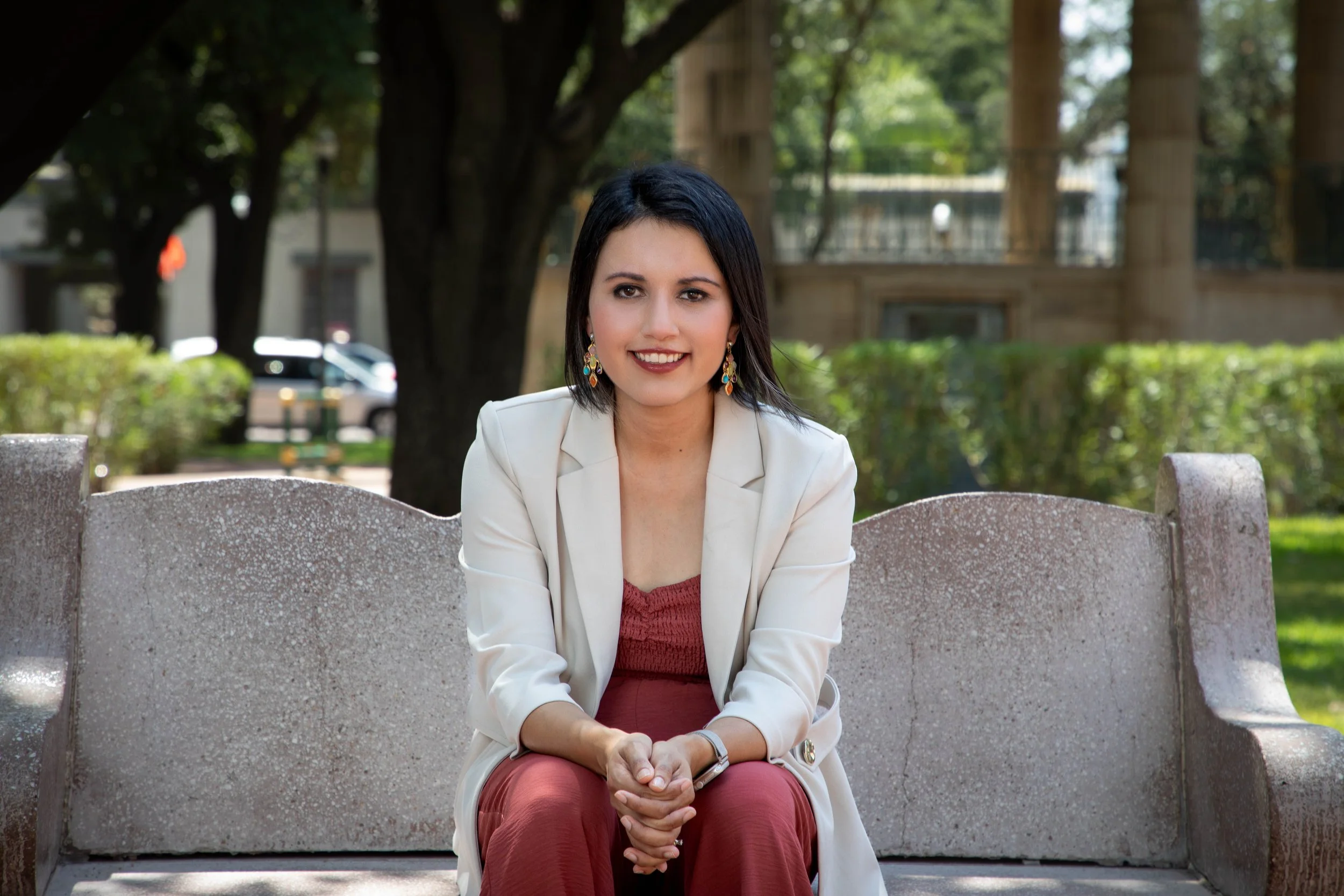 The therapist with black hair and earrings sitting on a stone bench outdoors, smiling at the camera. She is wearing a white blazer and a reddish-burgundy outfit, with trees and greenery in the background.