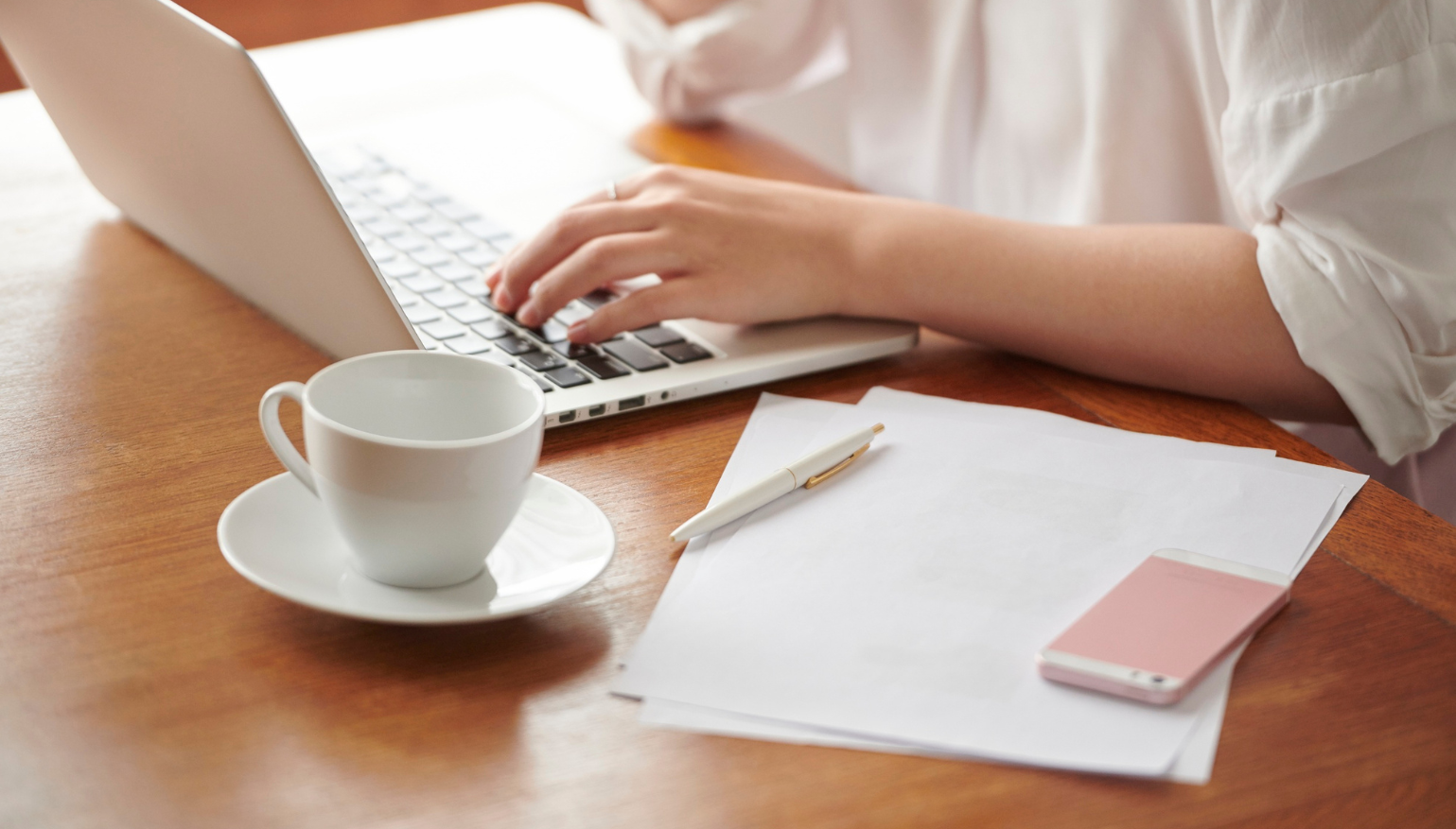 Person working on a laptop at a wooden desk with loose papers, a pink phone, a white pen, a white cup on a saucer, and a white shirt.