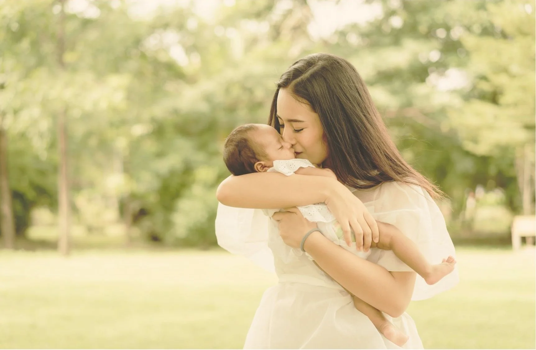 A young woman holding a baby outdoors in a park, both with closed eyes, sharing a kiss on the nose, surrounded by green trees and sunlight.