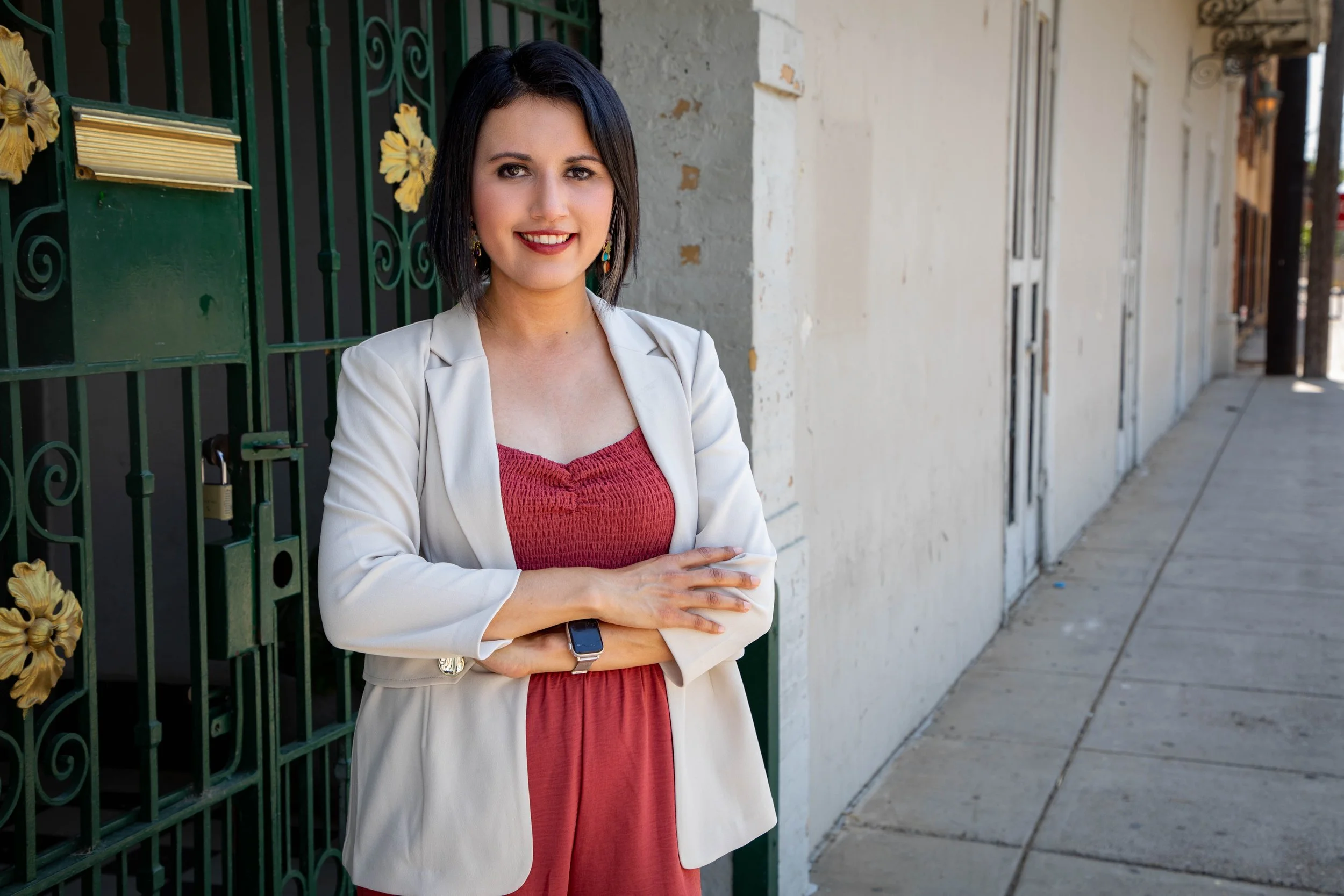 The therapist with black hair, wearing a white blazer over a red dress, standing outdoors against a green gate with gold decorations, smiling with her arms crossed, on a sidewalk with a building in the background.