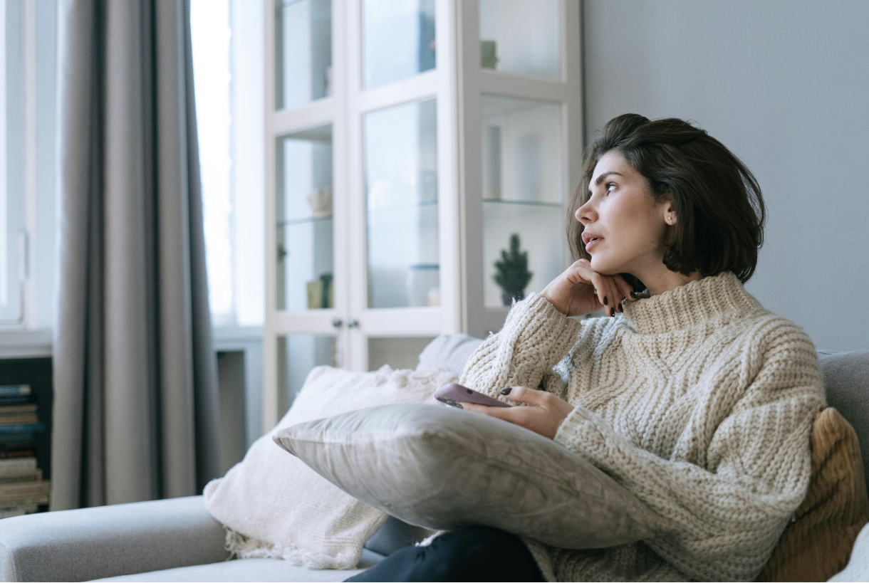 A woman with short dark hair sitting on a light-colored sofa, holding a smartphone in her left hand, with a pillow on her lap, in a cozy living room with a white cabinet and large windows with gray curtains in the background.