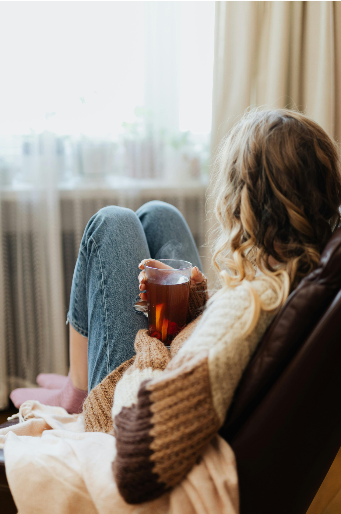A woman sitting in a chair, holding a steaming glass of tea, with legs raised and crossed, in a cozy indoor setting near a window with curtains.