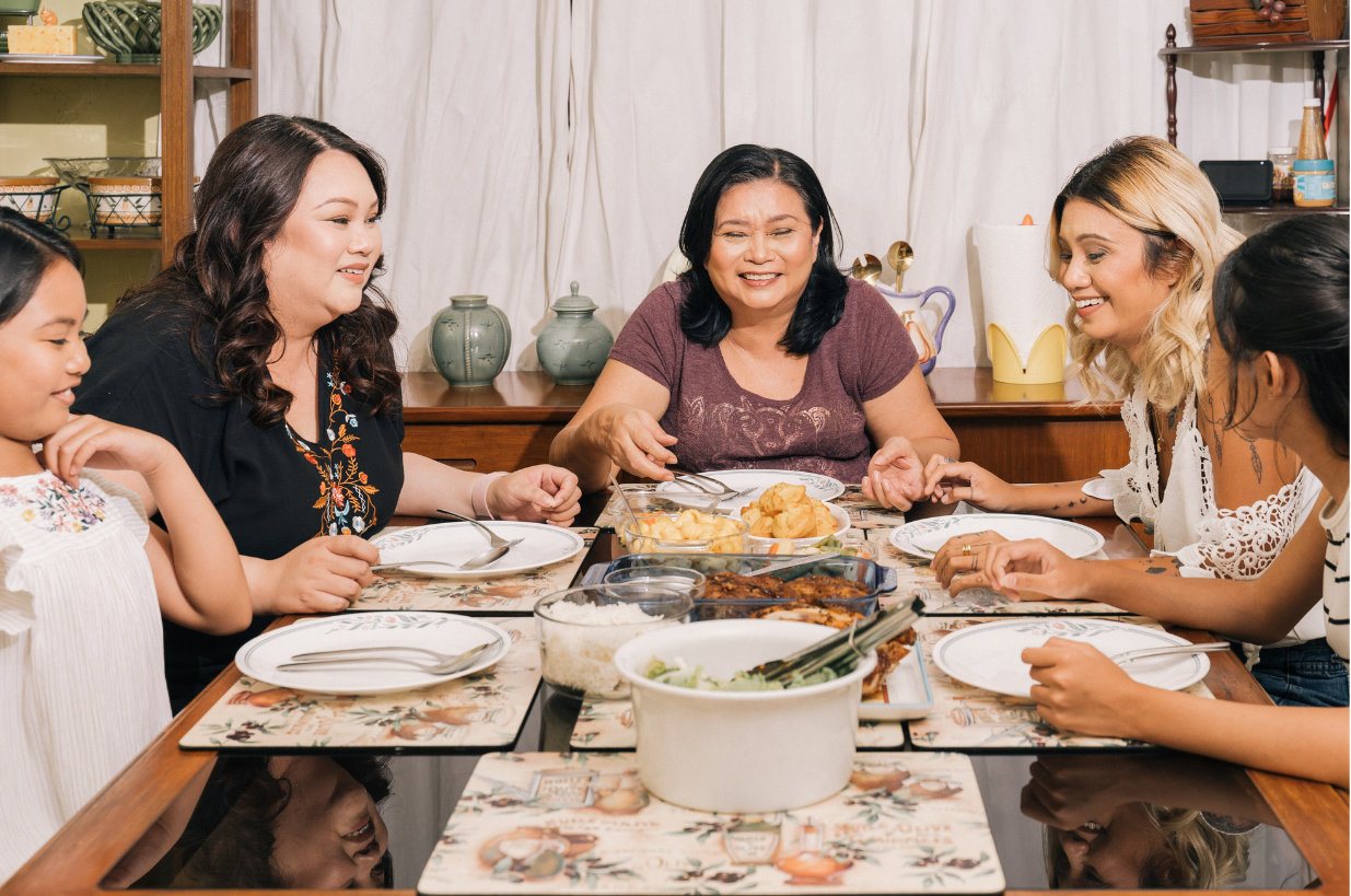 A diverse group of six women and one girl gathered around a dinner table enjoying a meal together. The table is covered with various dishes, including fried chicken, rice, and vegetables. They are smiling, engaging in conversation, and appear to be happy.