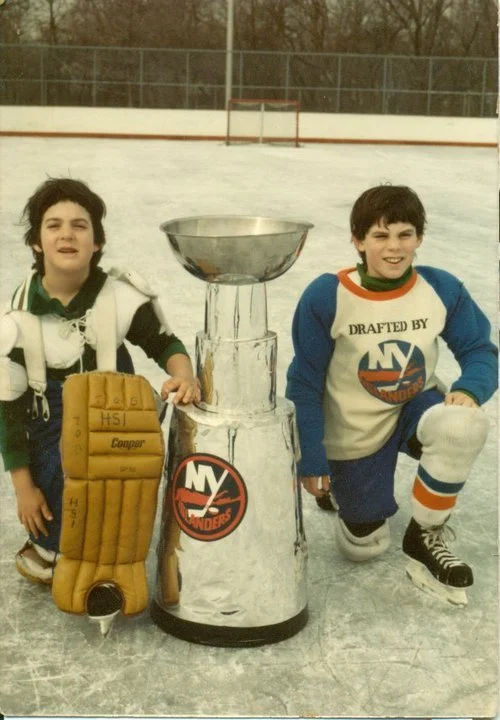 Two young boys in hockey gear kneeling on an ice rink next to a large replica trophy with the New York Islanders logo.