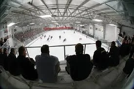 View of an indoor ice hockey game from the spectator stands showing players on the ice and spectators watching.