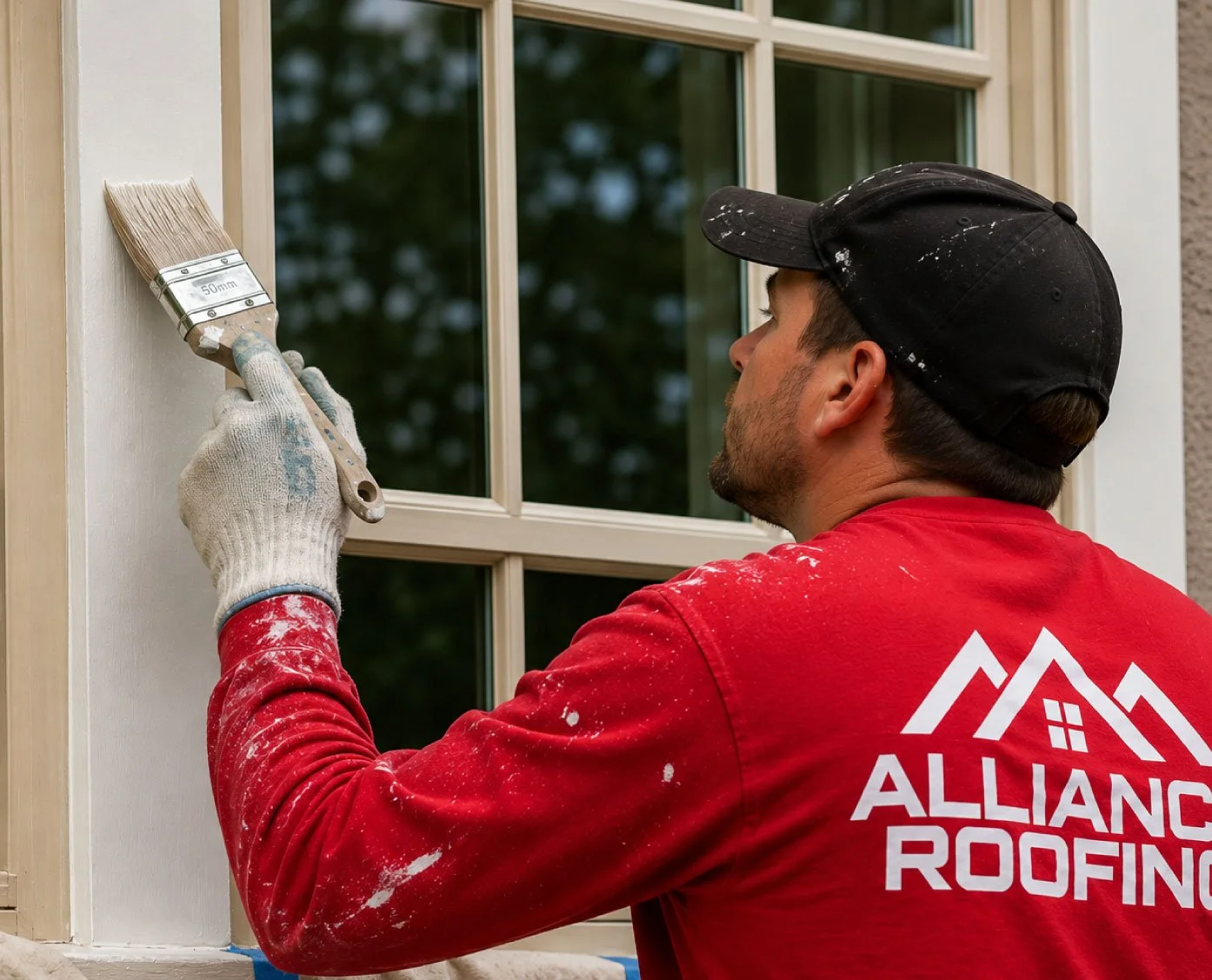 A man wearing a red shirt and a black cap is painting window trim with a brush on a white wall outside a house.