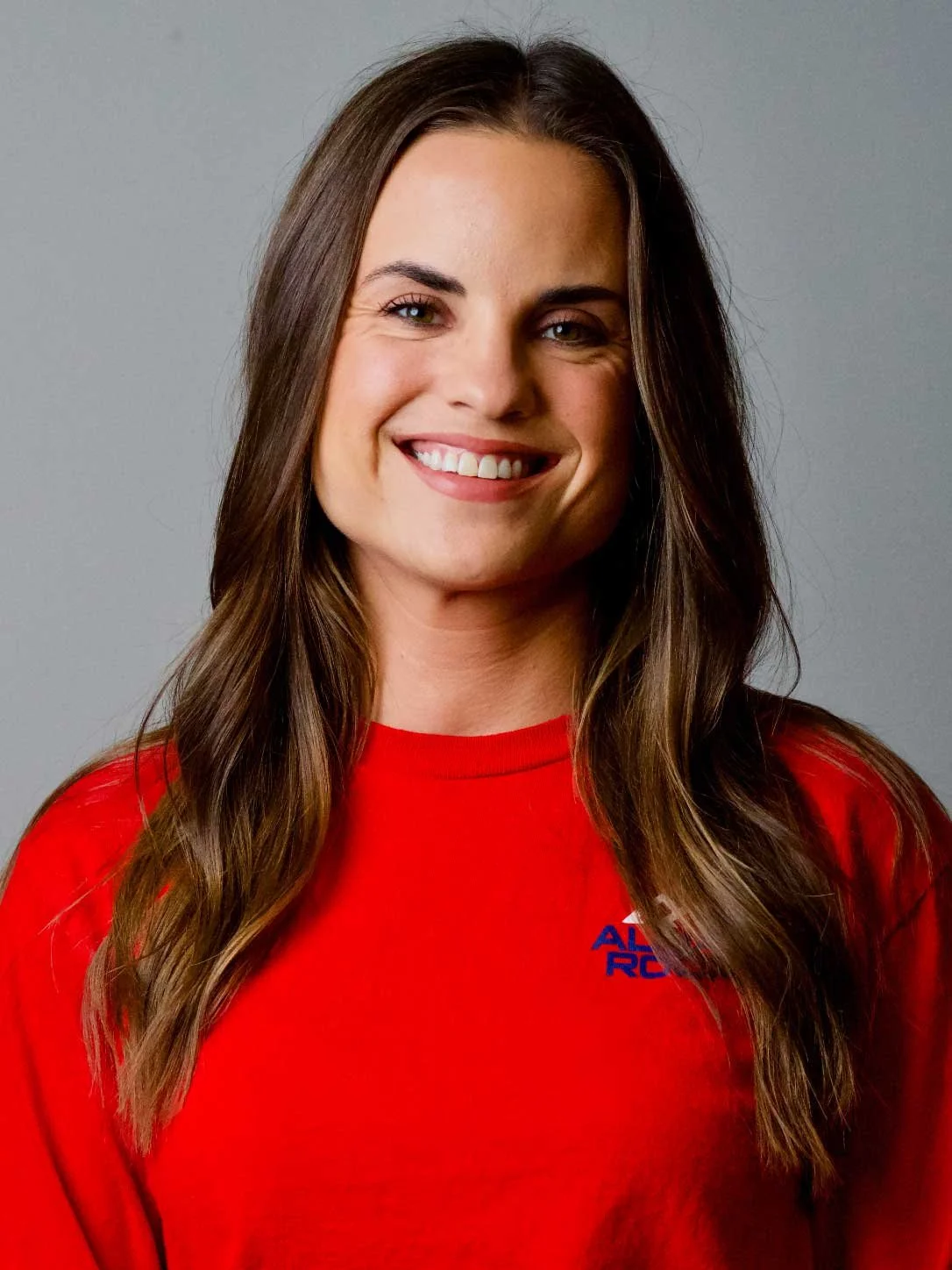 A woman with long brown hair wearing a red shirt, smiling, appearing to be in a casual setting.