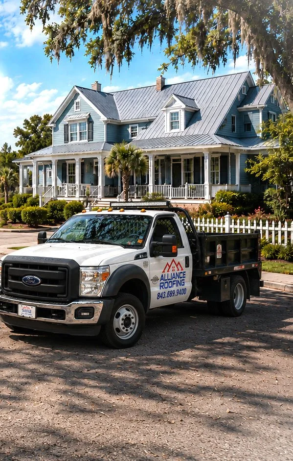 A large blue Victorian-style house with a metal roof, a porch, and multiple dormer windows, situated on a well-manicured lawn with a white picket fence. In front, there is a white service truck with 'Alliance Roofing' logo parked on the street.