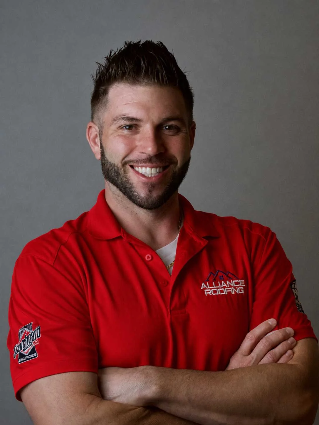 A man with a beard and short brown hair, smiling with arms crossed, wearing a red polo shirt with an 'Alliance Roofing' logo on the front and another logo on the sleeve, standing against a gray background.