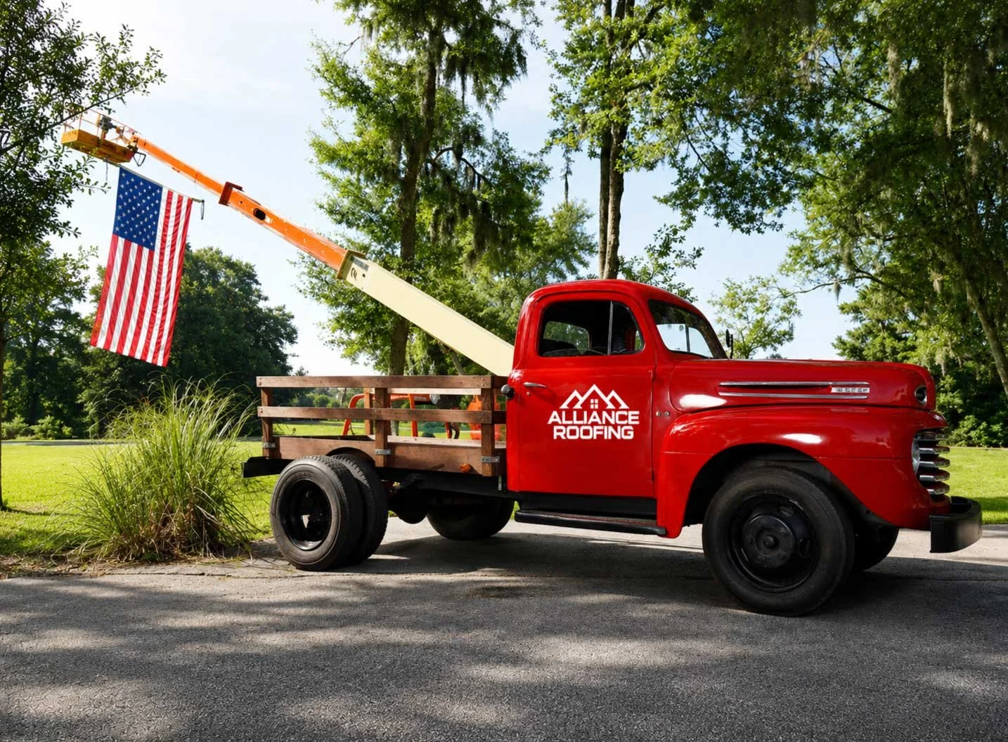 Red vintage pickup truck with 'Alliance Roofing' logo parked on a paved road, hanging American flag from an extendable arm, surrounded by green trees and grass.