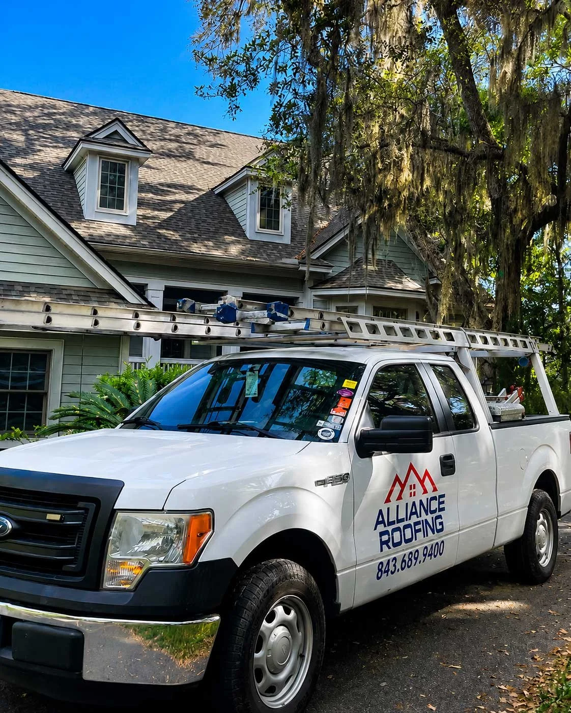 A white service truck for Alliance Roofing parked in front of a house with a ladder on top. The house has multiple dormer windows, light green siding, and is surrounded by trees.
