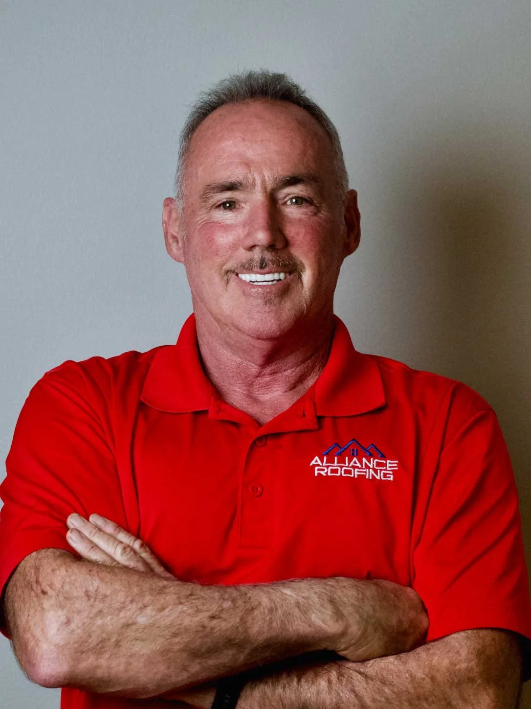 A middle-aged man with short gray hair and a mustache, smiling, wearing a red polo shirt with the words 'Alliance Roofing' and a roof graphic embroidered on it. He is standing with his arms crossed against a gray wall background.