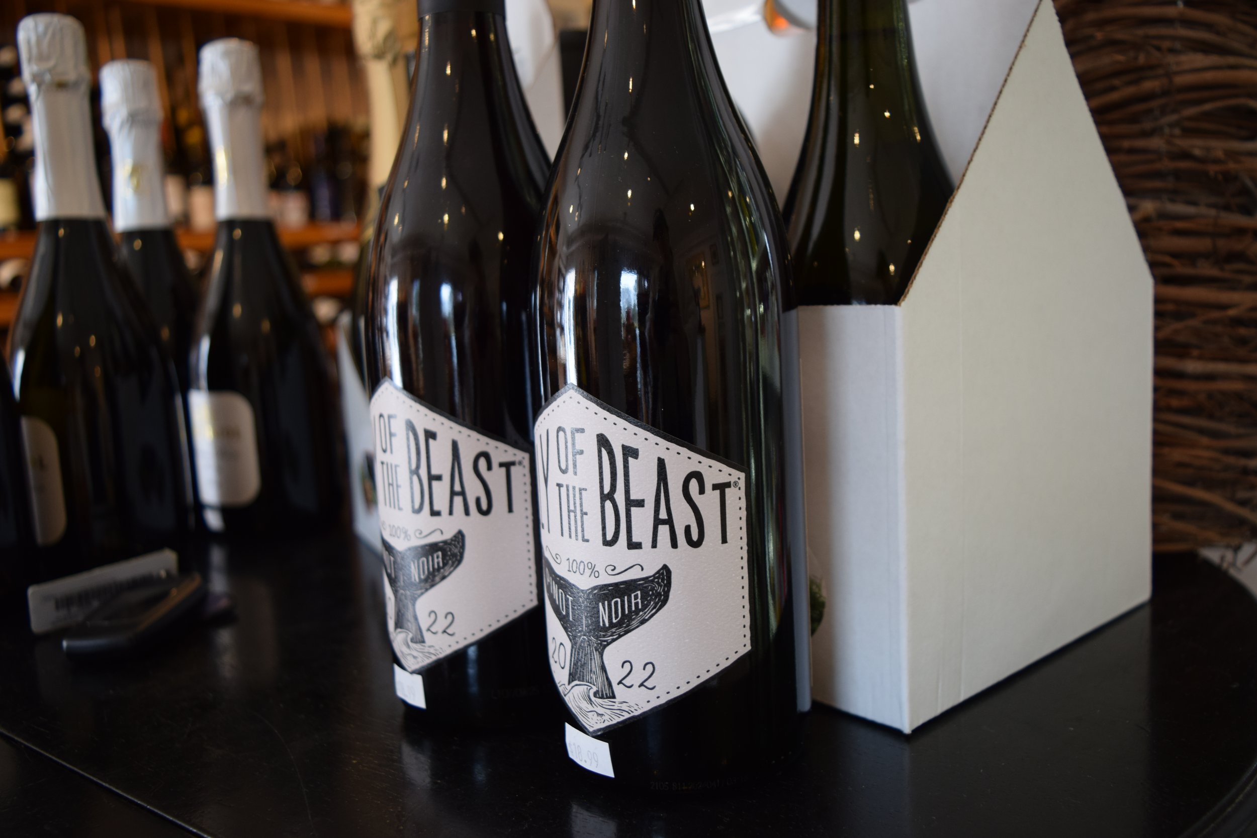 Several bottles of black beer with white labels and black caps are displayed on a black surface in a store. The labels read "Of The Beast Noir" and indicate 100% dark stout beer priced at $10.99.