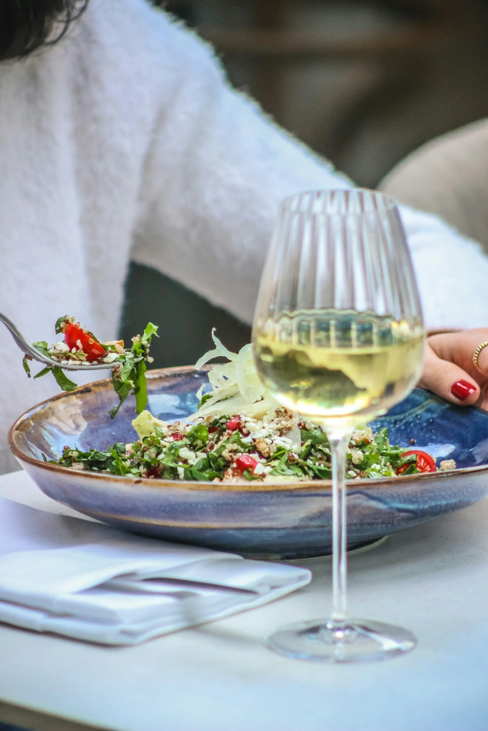A close-up of a plated salad with various greens, pomegranate seeds, and shredded cheese, accompanied by a glass of white wine on a table set with white napkins.