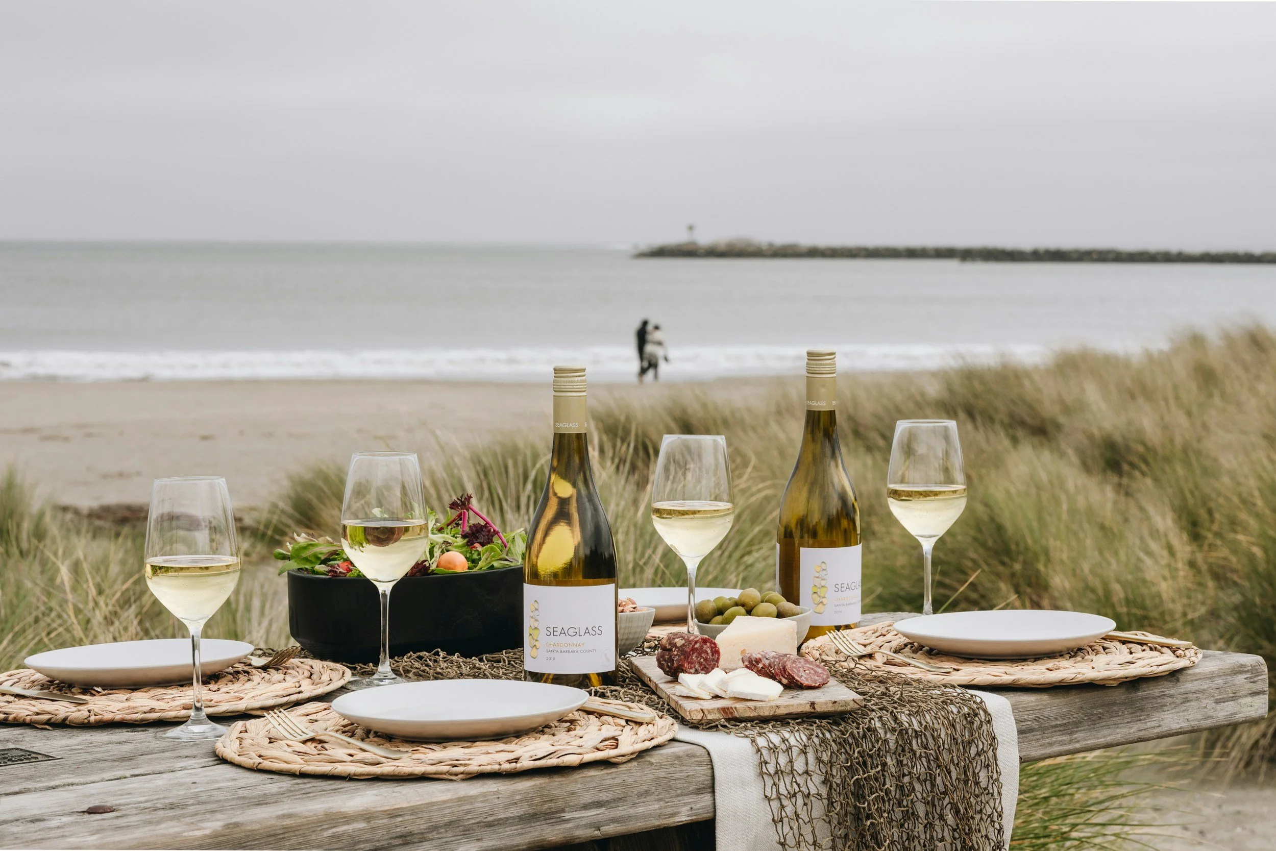 A beachside dining setup with wine glasses, bottles of white wine, cheese, and charcuterie, overlooking the ocean with a rocky pier and overcast sky in the background.