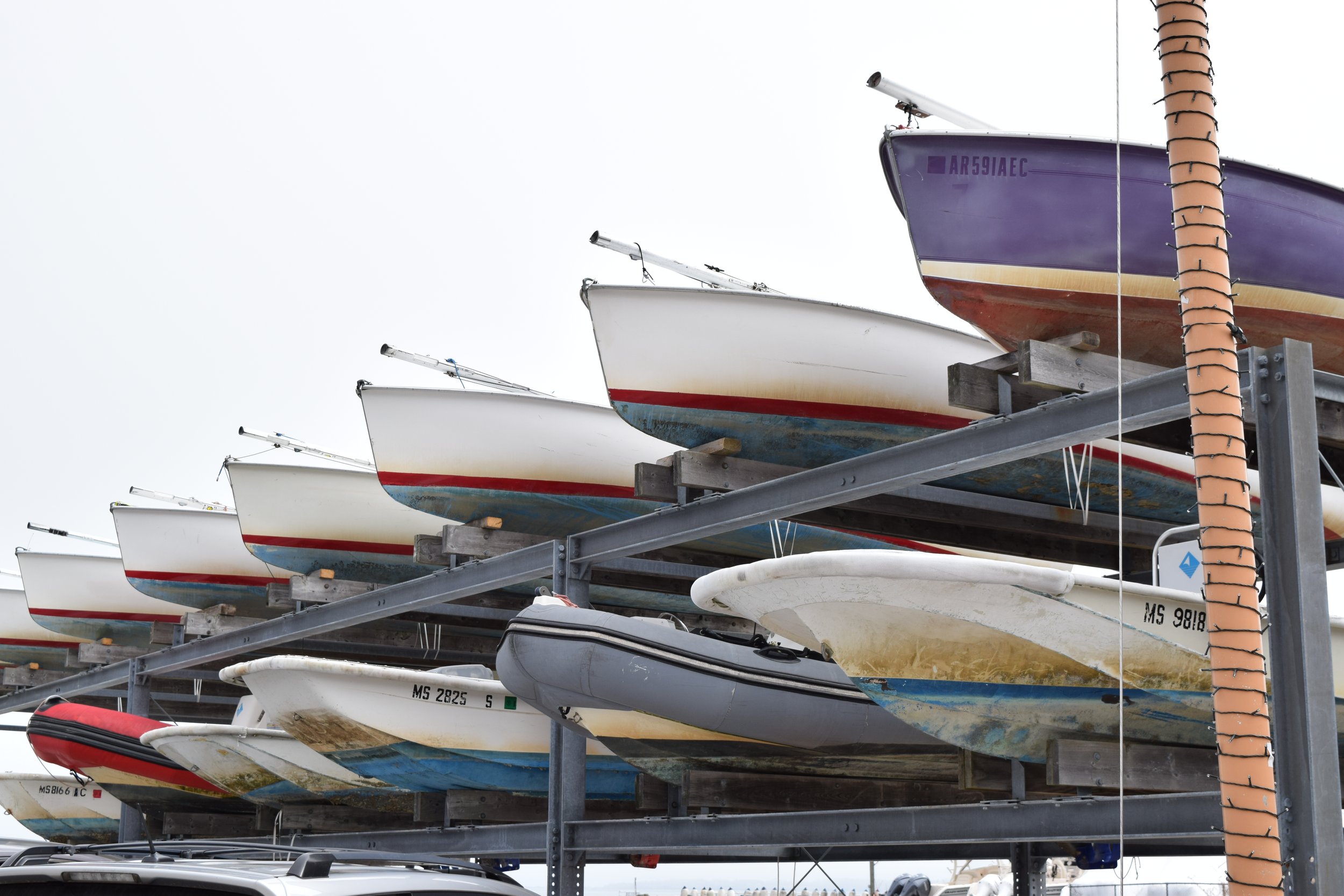 Multiple boats stored on a metal boat rack, with some showing its hulls and others with parts of their bottoms visible, against an overcast sky.