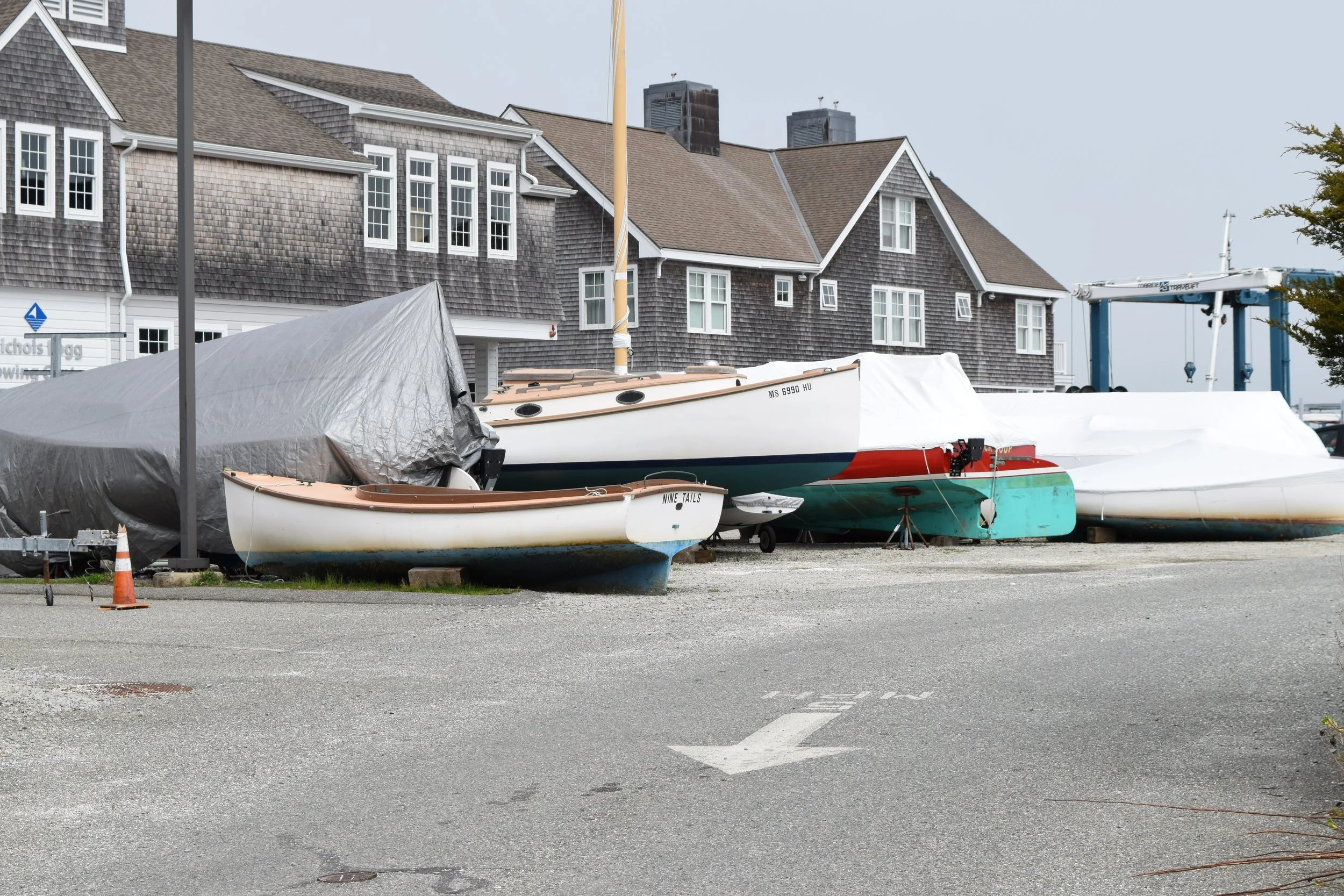 Several boats covered with tarps and boat covers, parked on a gravel lot near houses with shingle siding, some automotive equipment, and trees in the background.