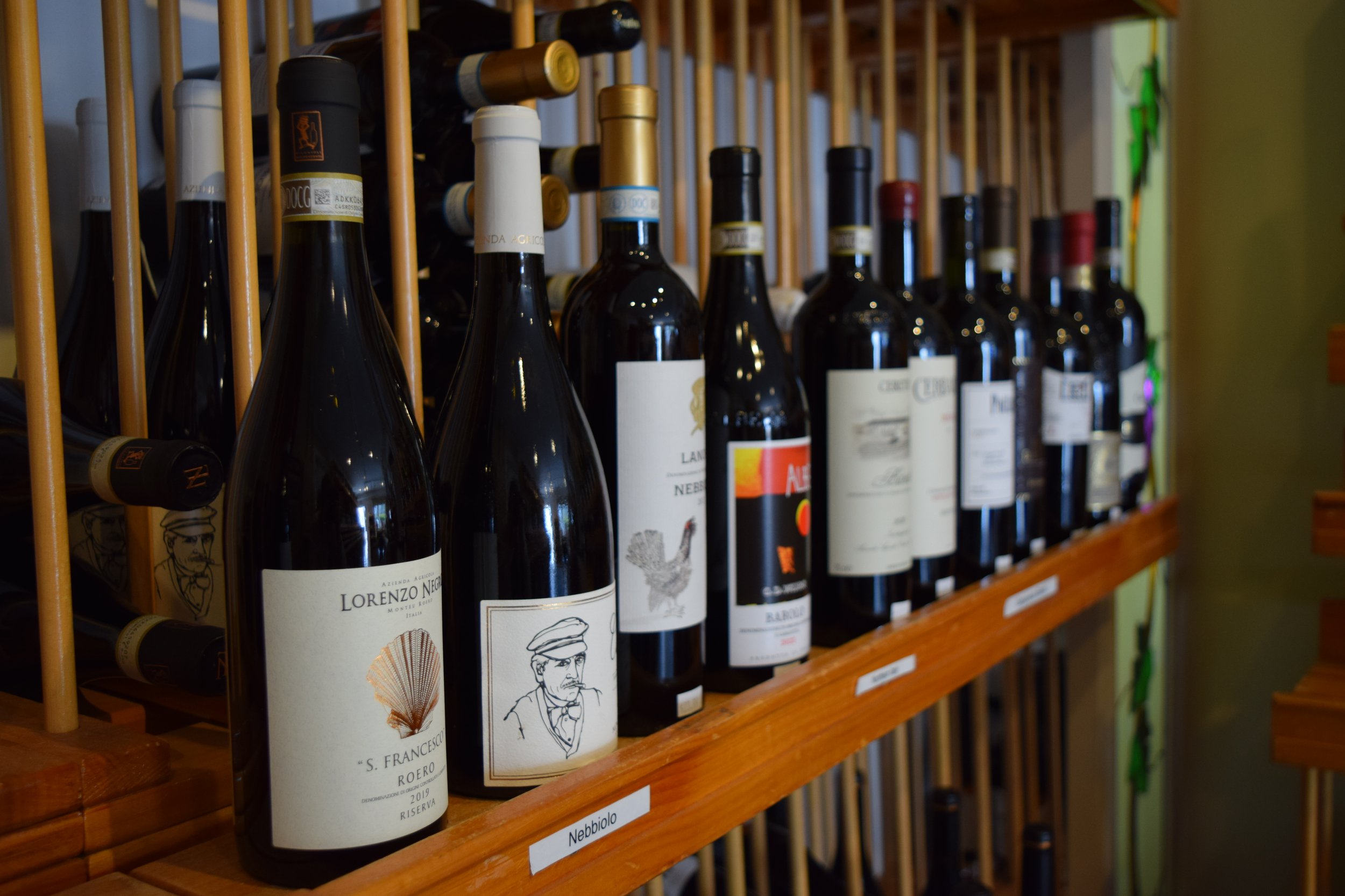 Rows of wine bottles on a wooden wine rack with labels visible, including names like Lorenzo Negri and S. Francesco. The bottles are stored horizontally and have various label designs.