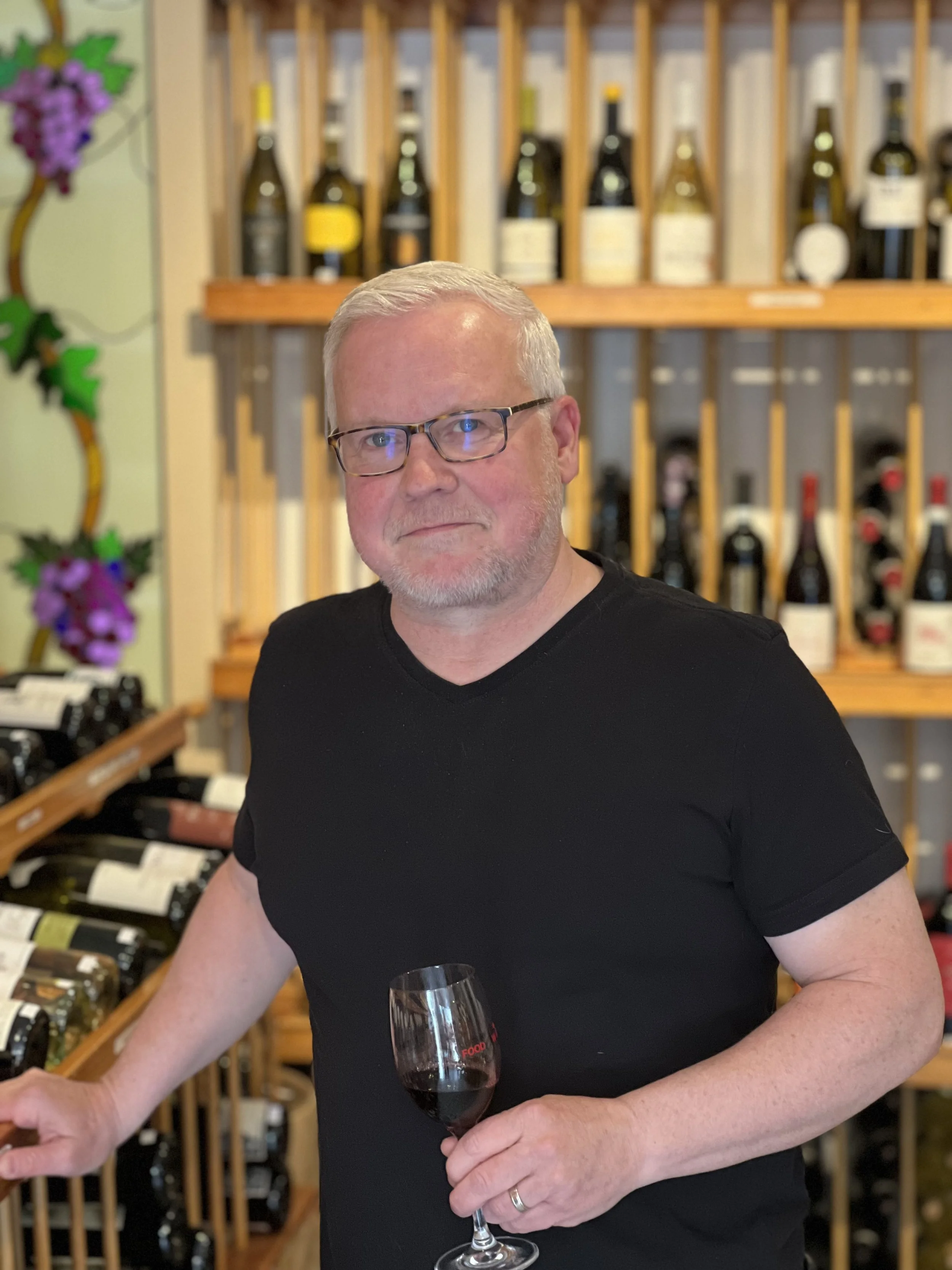 A man in a black t-shirt and glasses holding a glass of red wine in a wine shop with bottles displayed on wooden racks behind him.
