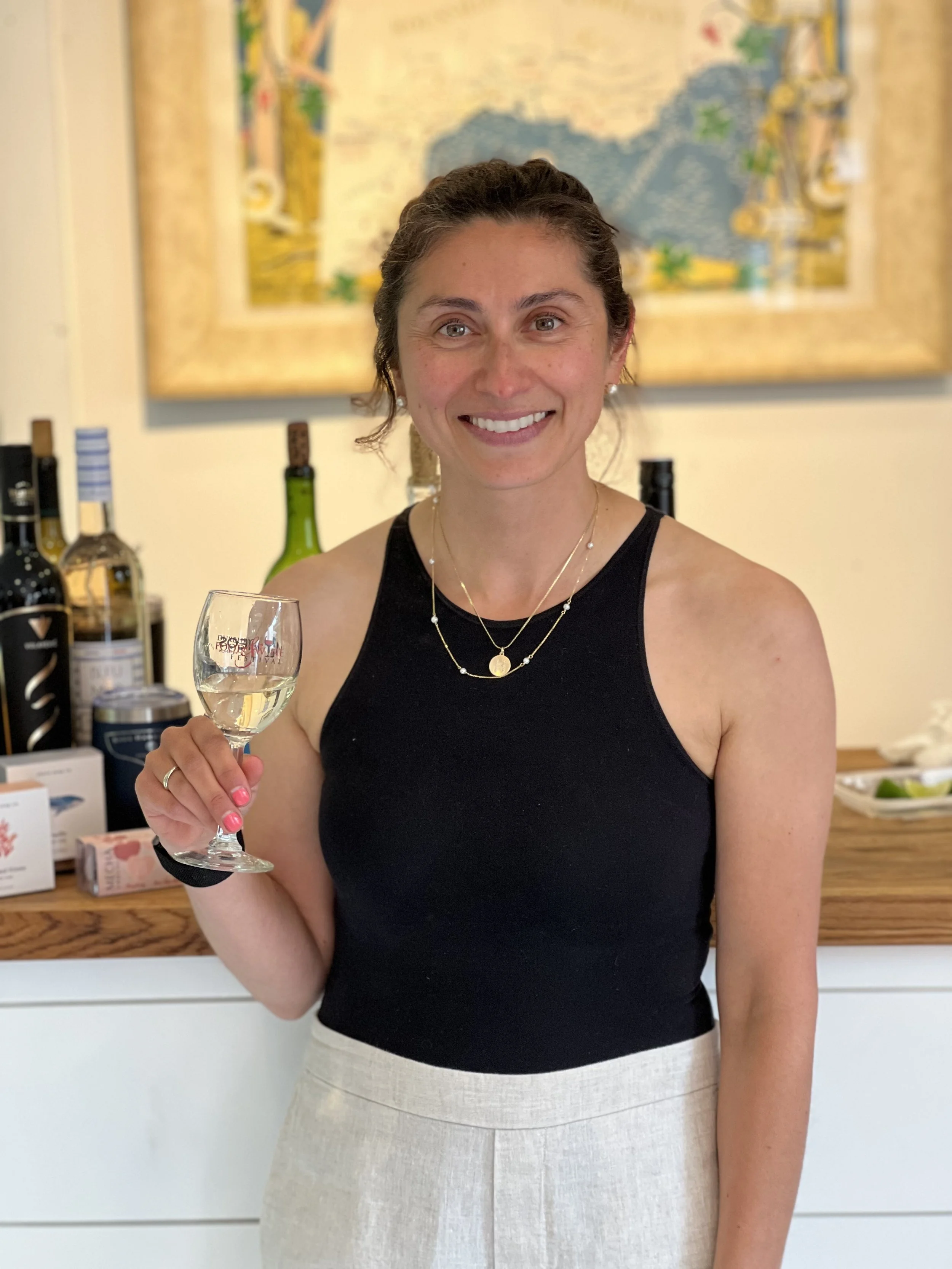 A woman with dark hair smiling and holding a wine glass, standing in front of a wall with bottles and a framed map.
