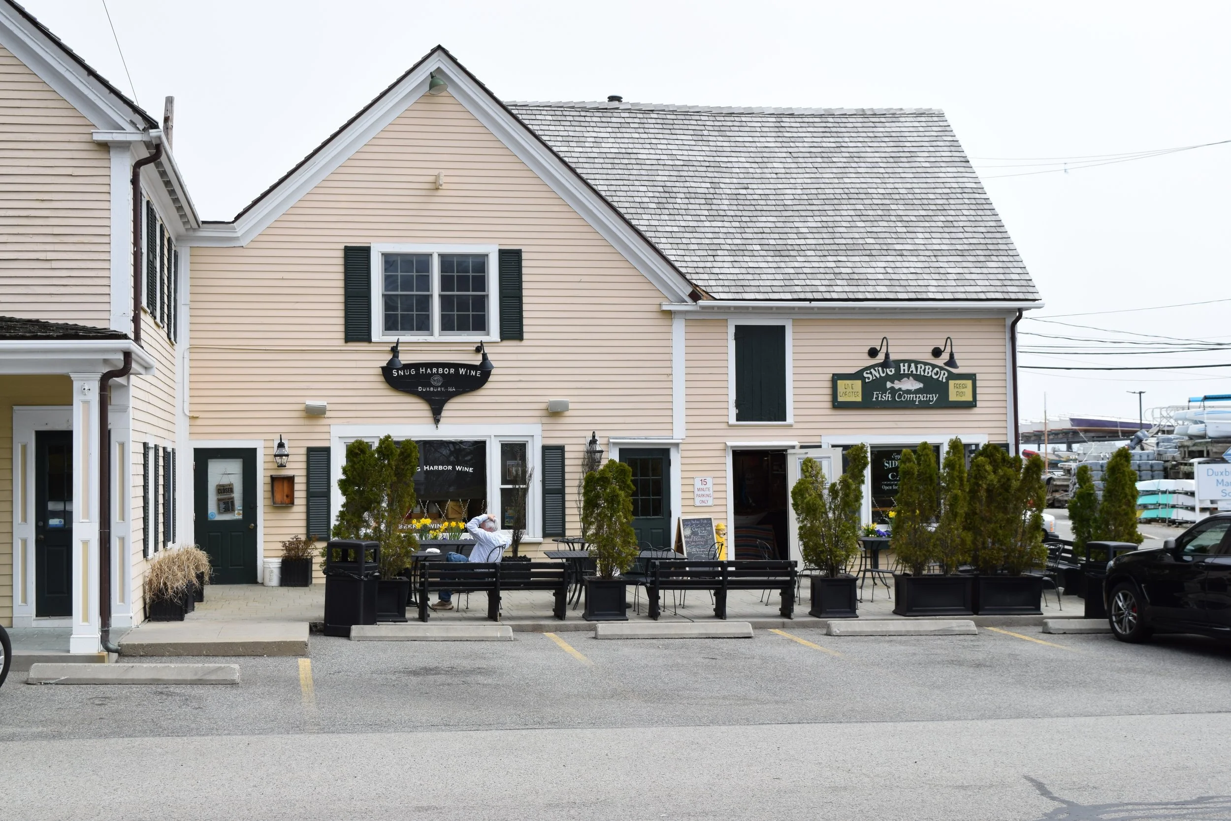 A beige building with black shutters and a sign that reads 'Snug Harbor Wine' and 'Snug Harbor Fish Company.' There are outdoor tables and chairs in front of the building with a few people seated. The parking lot has trees in black planters and some 