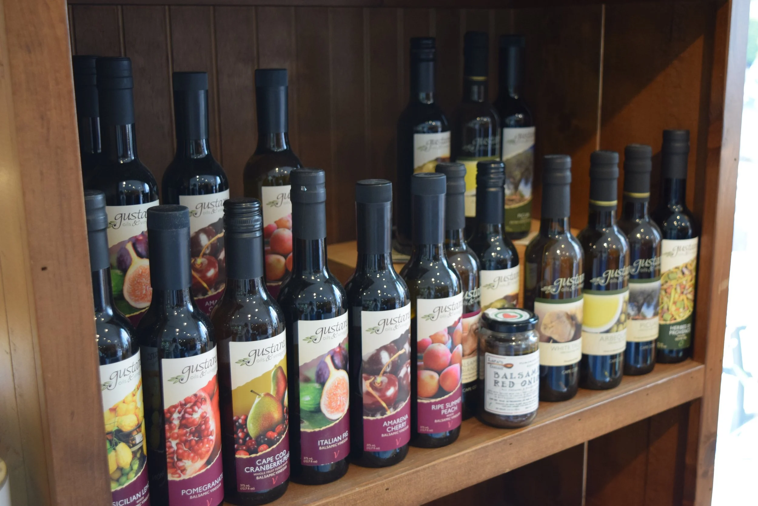 A wooden shelf displaying bottles of flavored balsamic vinegar with colorful fruit labels and a few spice jars.