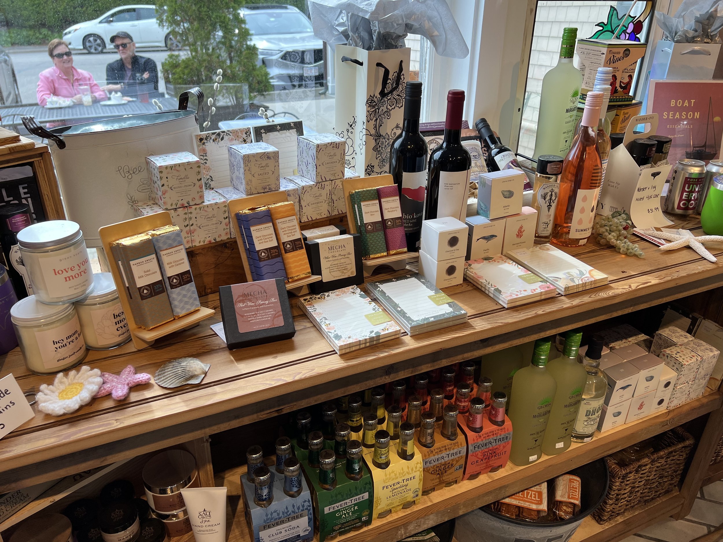 A display table with assorted bottles of wine, boxes of chocolates, and various beverages in a store. Outside the window, two people are sitting at a table, visible through the window.