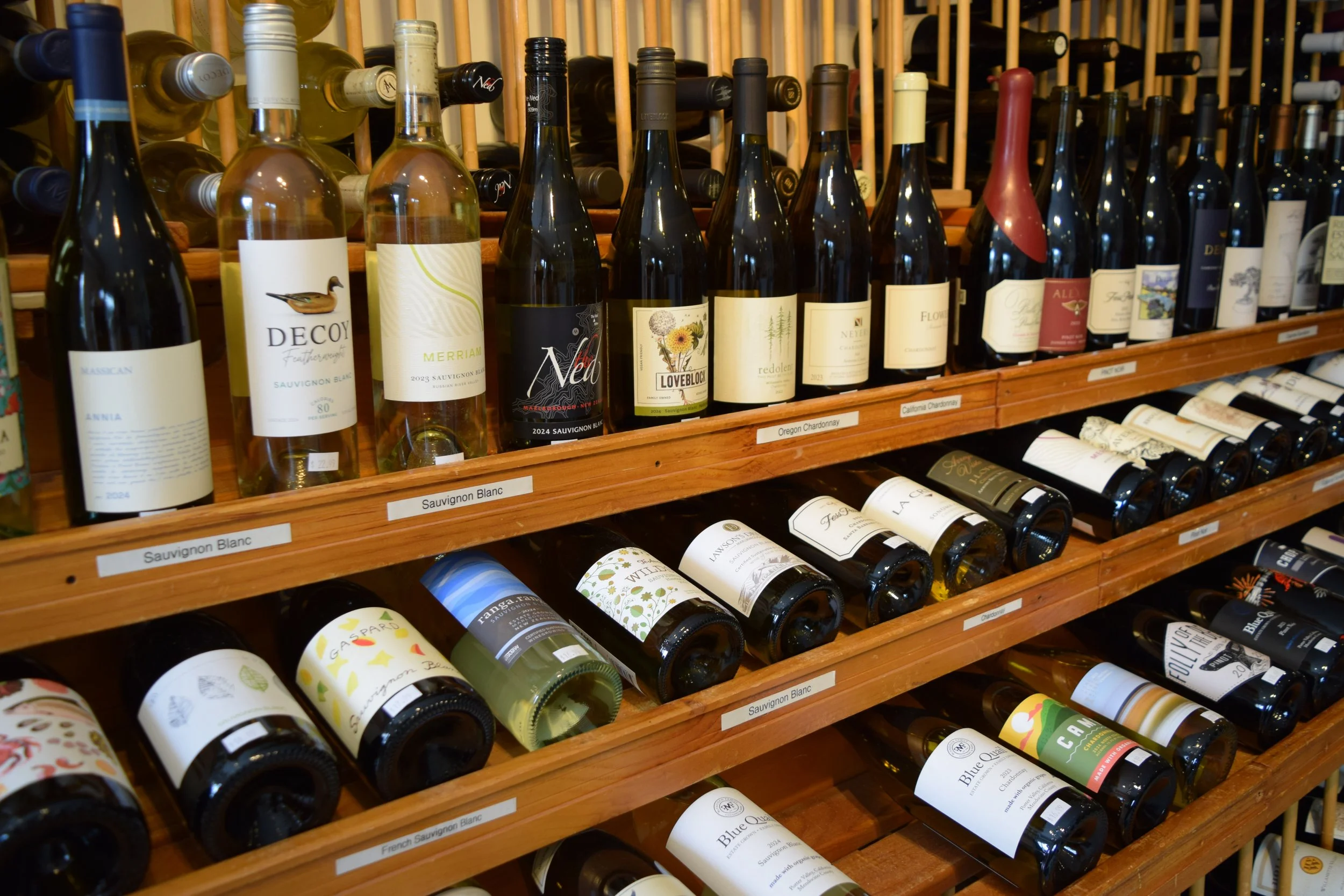 A variety of wine bottles displayed on wooden shelves, labeled with different wine types such as Sauvignon Blanc and Chardonnay.