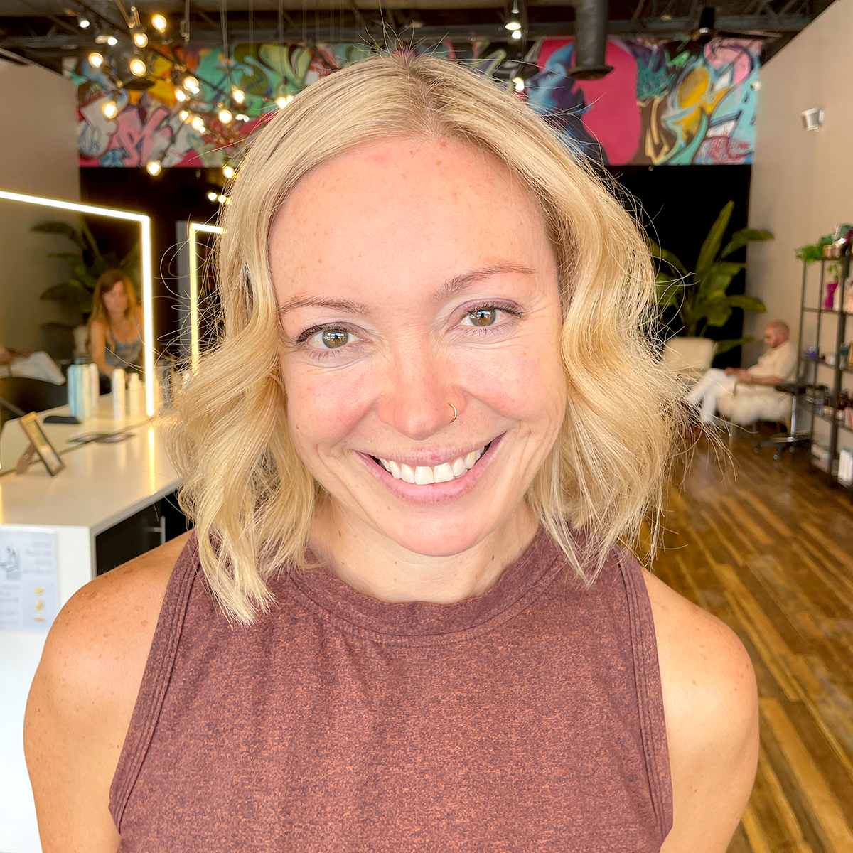 A smiling woman with wavy blonde hair and a nose piercing, wearing a sleeveless maroon top, in a modern salon with colorful graffiti art on the walls, large mirrors, and a wooden floor.
