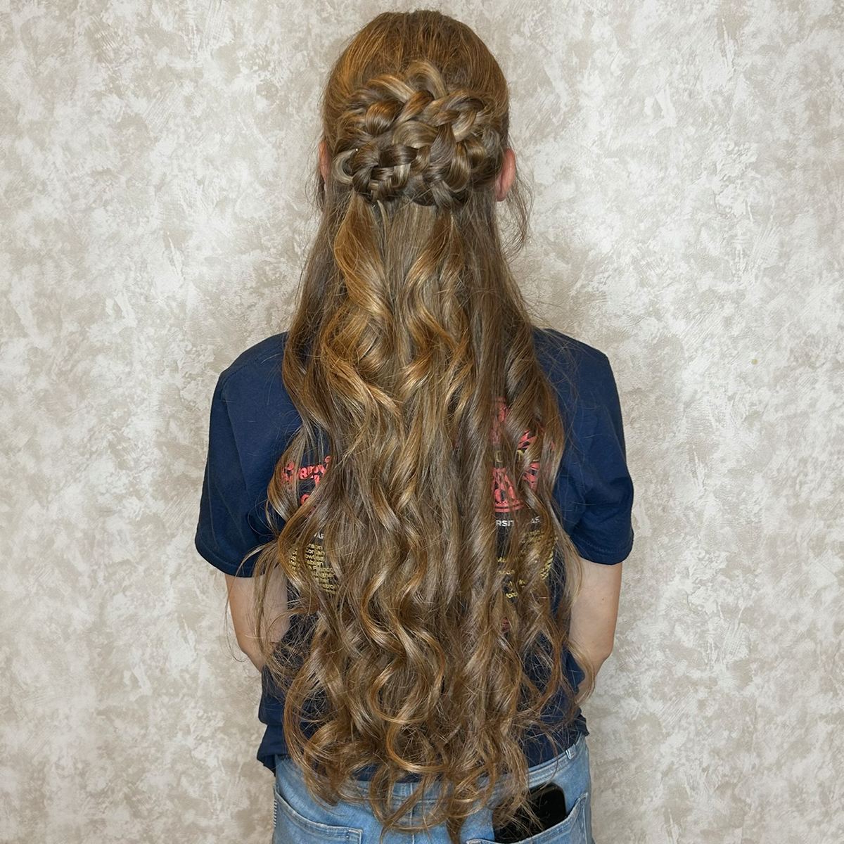 Back view of a woman with long, curly, light brown hair styled with a braid across the top and loose curls flowing down.