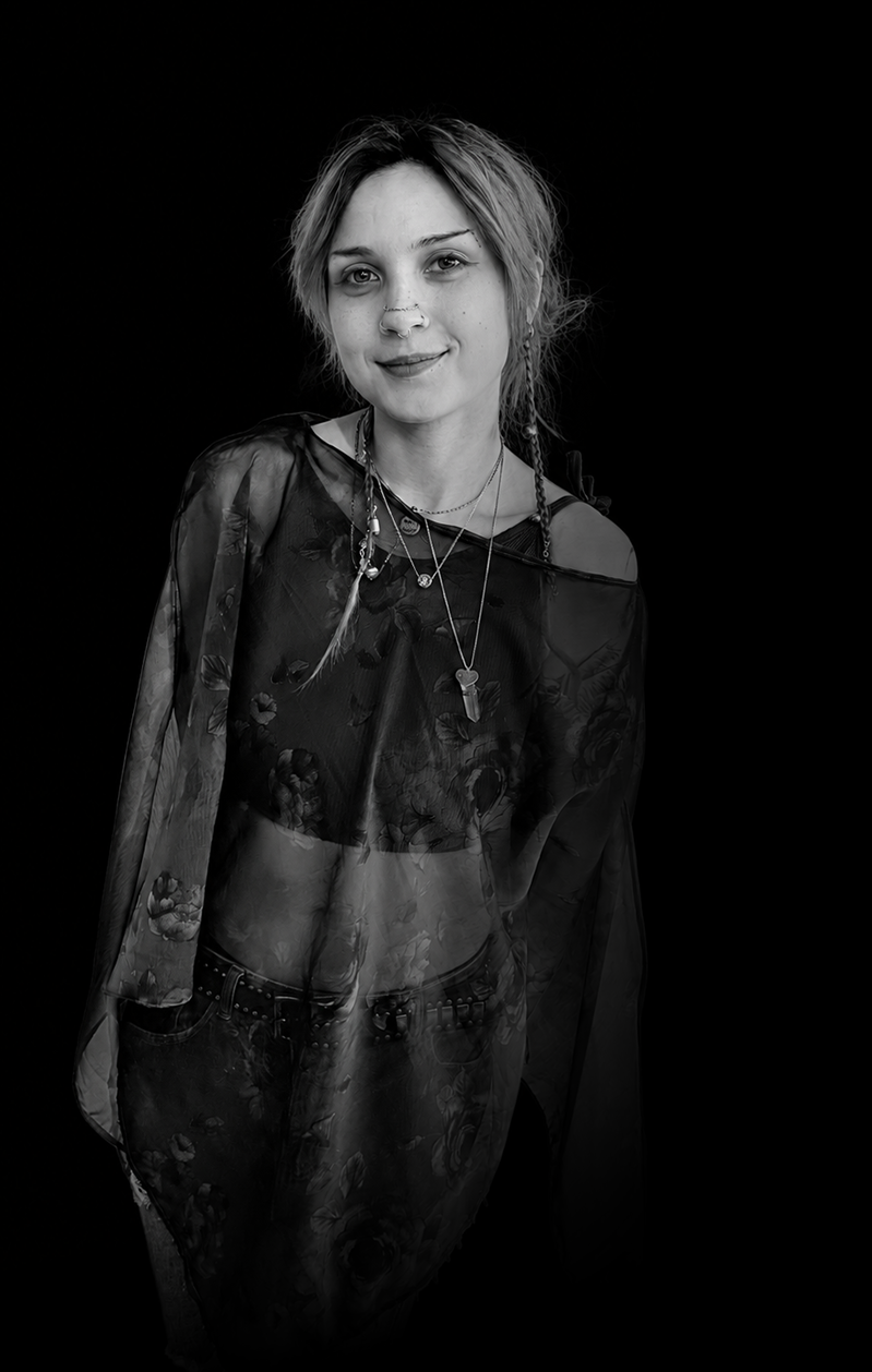 Black and white portrait of a young woman with short, tousled hair, wearing layered necklaces and a semi-sheer floral top with matching pants, standing against a dark background.