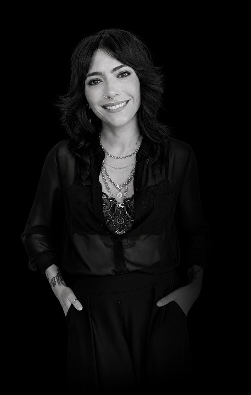 Black and white portrait of a woman with shoulder-length wavy hair, wearing layered necklaces, a nose ring, and a dark semi-sheer blouse, smiling against a black background.