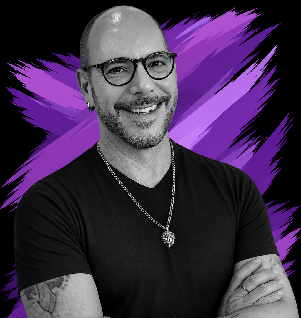 A smiling man with glasses, a beard, and a tattoo on his arm, wearing a black t-shirt and a chain necklace with a lion pendant, standing against a black and purple brushstroke background.